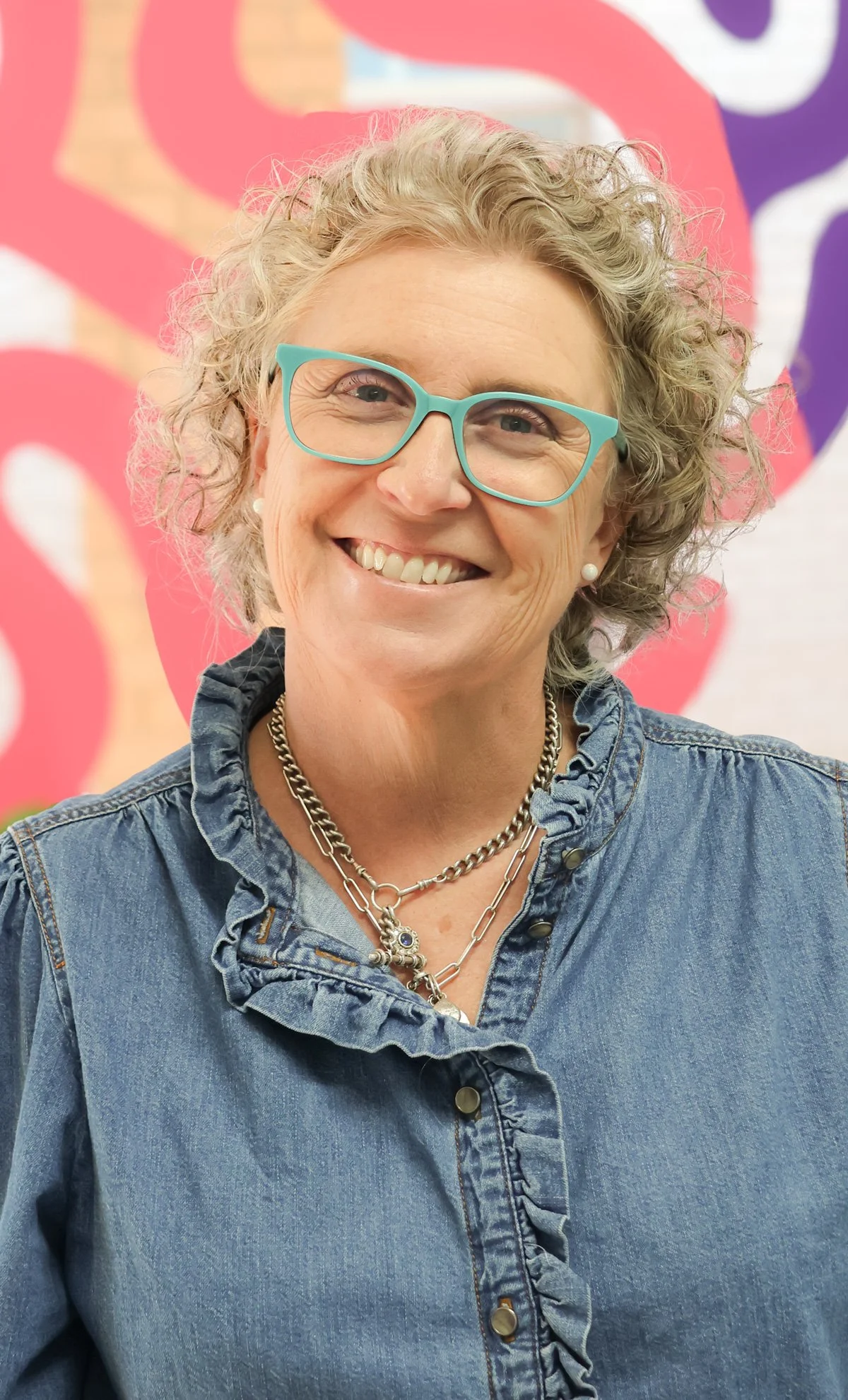 Fi Waters stands in front of the Messy Minds logo. She is smiling and wearing glasses.