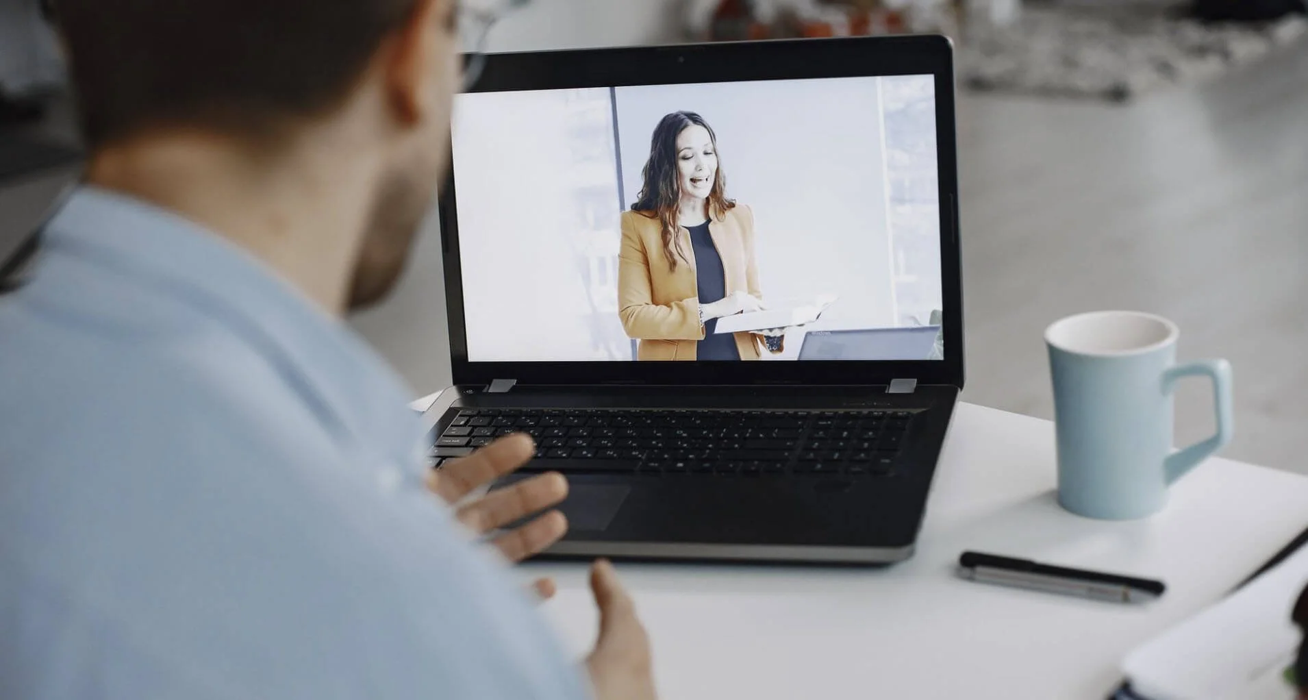 Person on video call with woman holding papers, sitting at a white table with a mug, pen, and notebook.