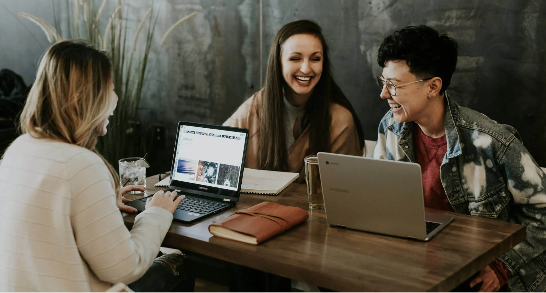 Three women sitting at a wooden table, smiling and laughing, with laptops and notebooks in front of them