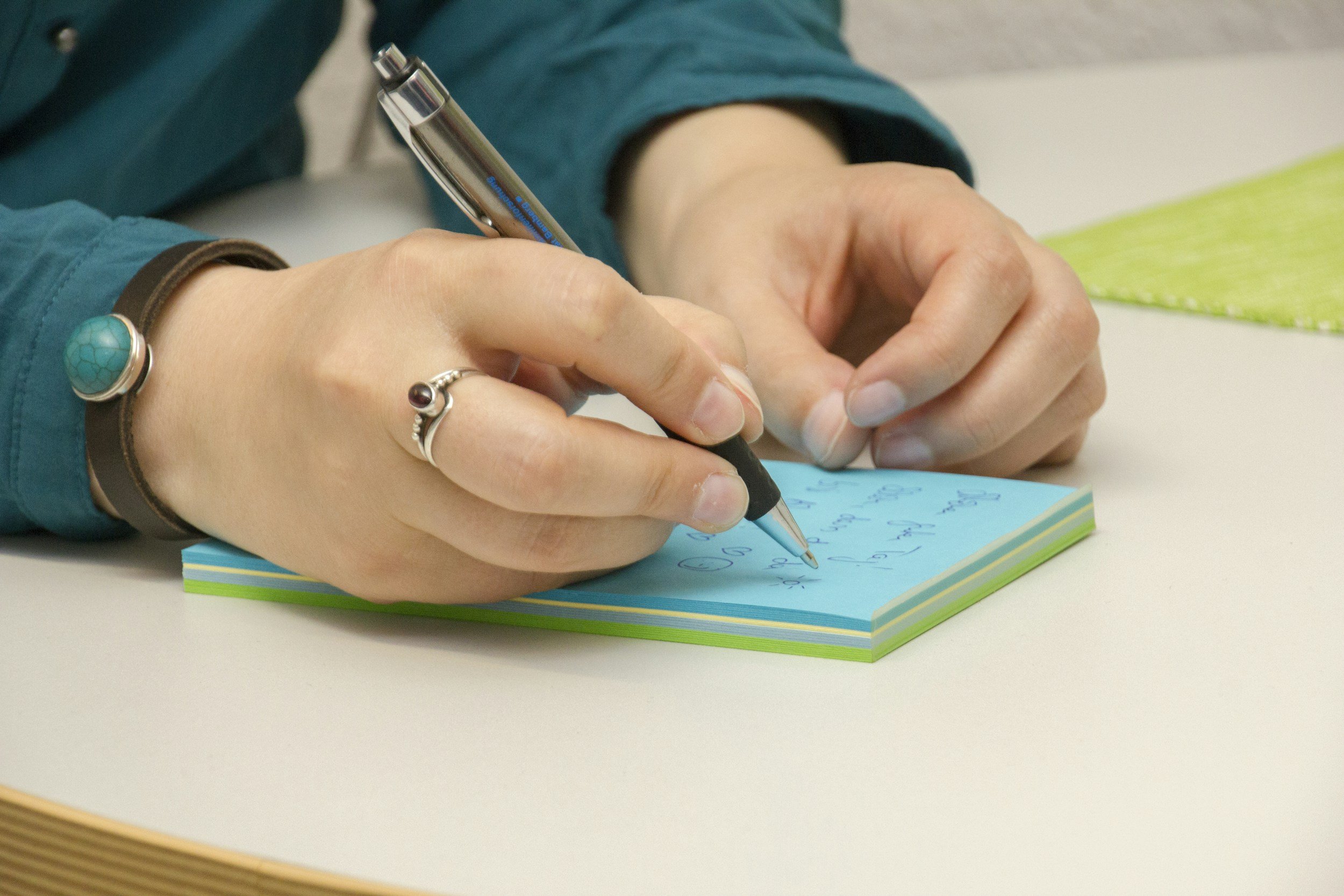 A person wearing a teal shirt writes notes on a blue notepad with a black pen. The person wears a ring with a large turquoise stone and a silver ring with a small red stone.