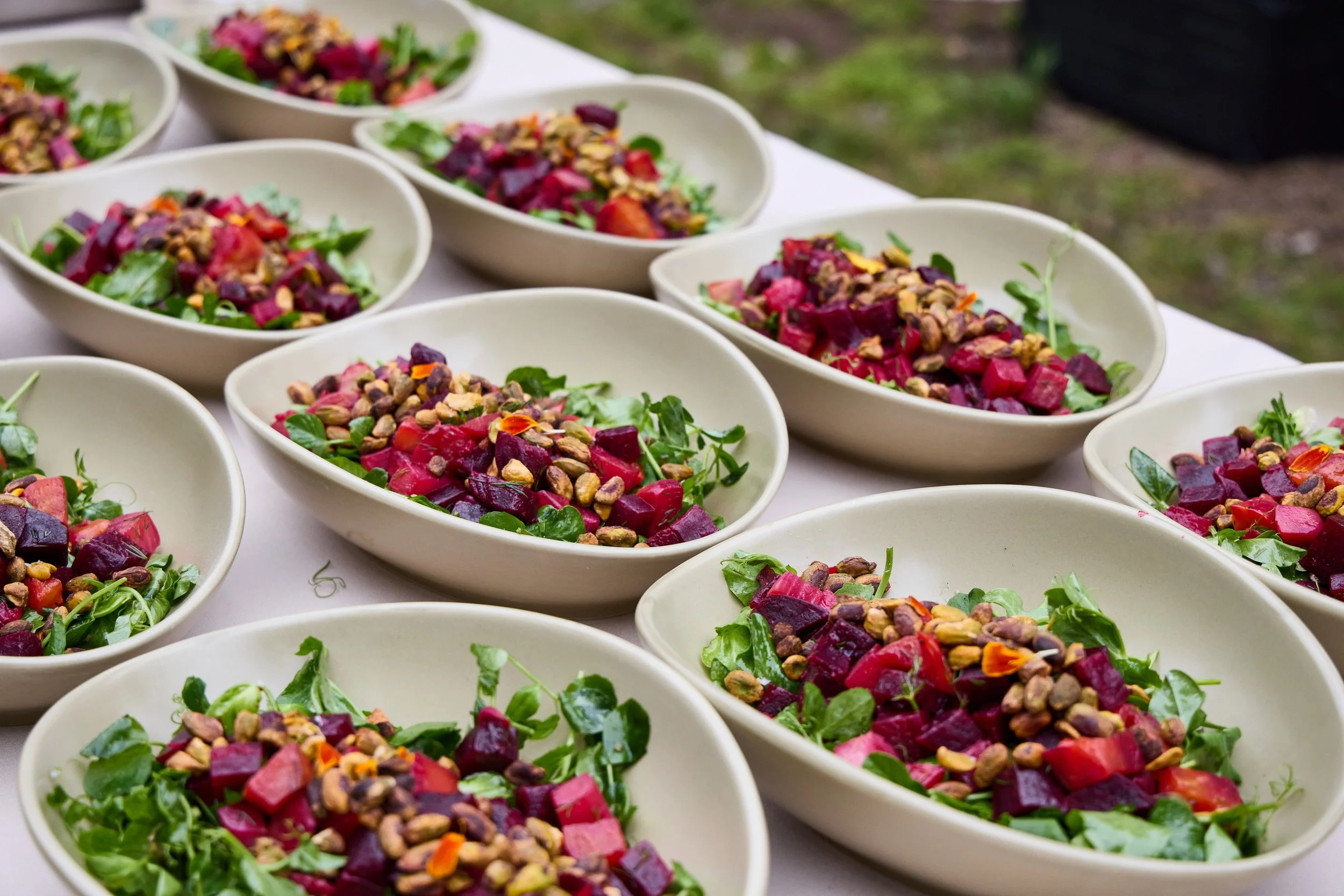 Several white oval bowls filled with colorful salads containing farm-to-table greens, beets, vegetables, and topped with toasted nuts are arranged on a white surface outdoors for Debonair Events client.