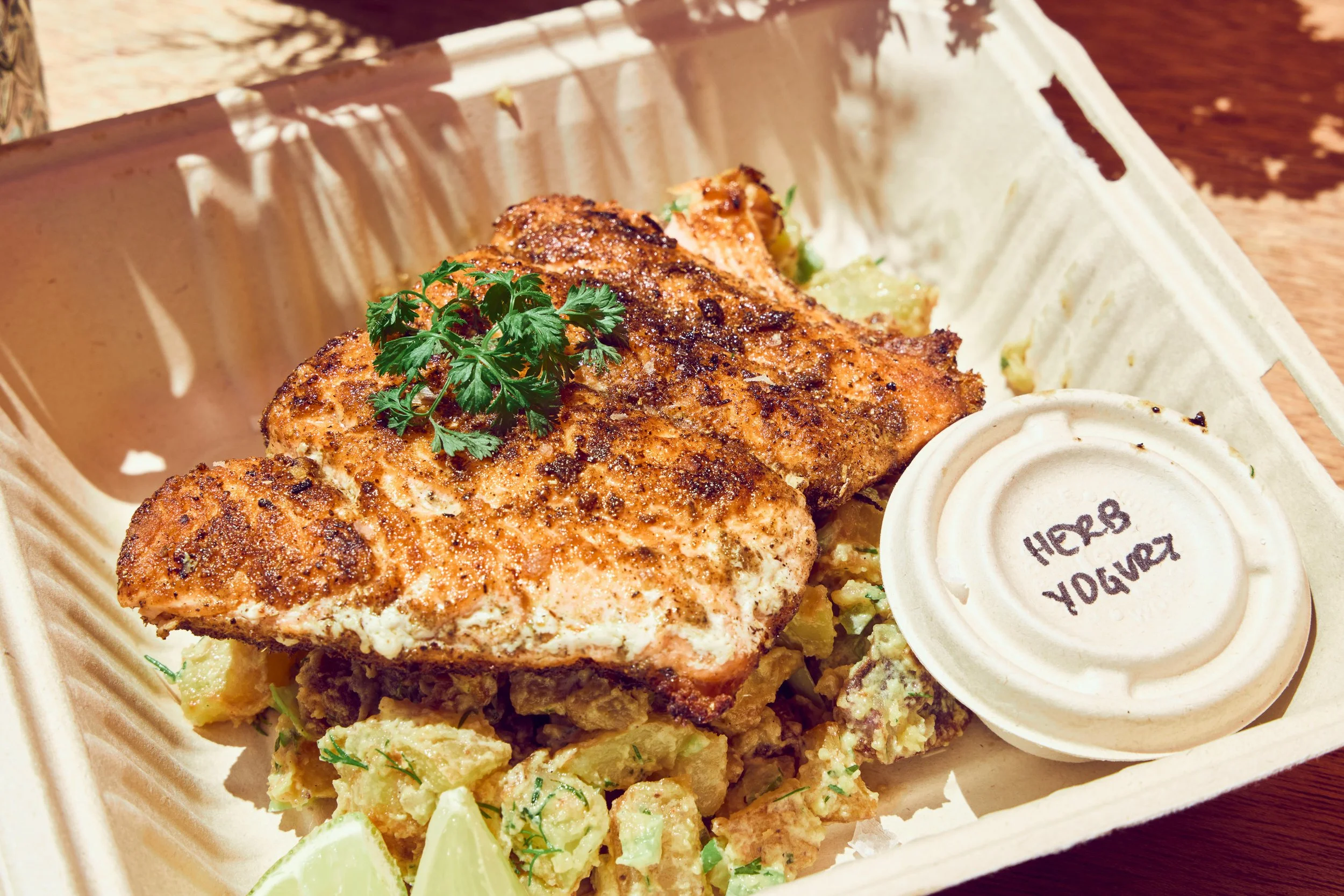 Grilled Bolinas salmon garnished with parsley served with summer potato salad, lime wedges, and a small cup of dipping sauce on a takeout tray.
