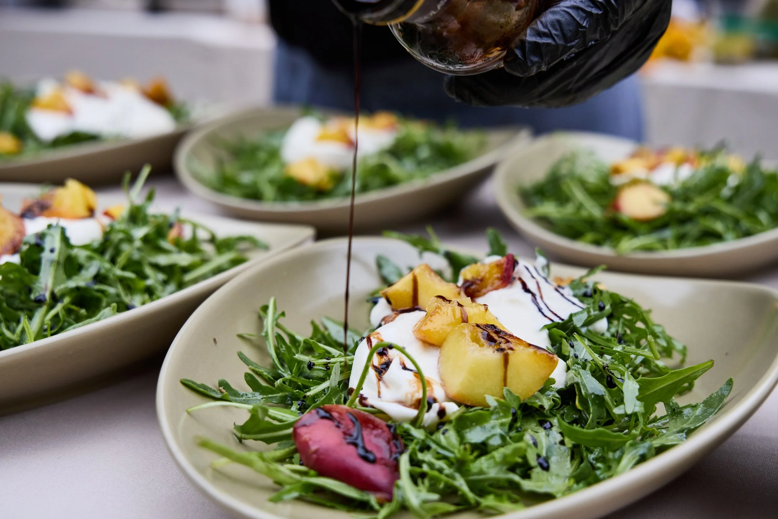 Salad bowls with arugula, grilled peach slices, and drizzled balsamic glaze, with balsamic vinegar being poured over one of the salads.