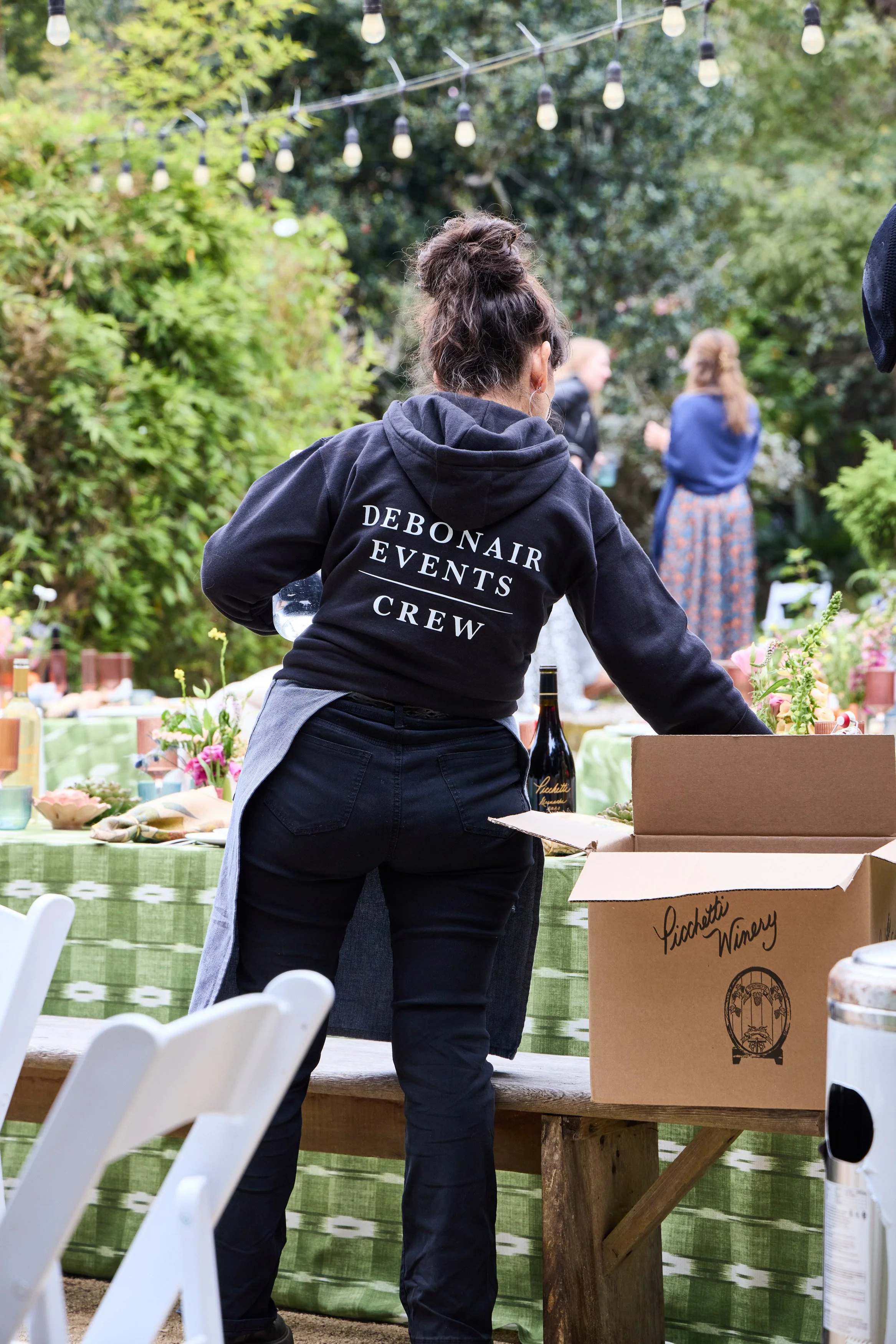 A Debonair Events crew member wearing a black hoodie with 'DeBonaire Events Crew' printed on the back, preparing the table at an outdoor wedding, with trees and string lights overhead, and guests in the background.