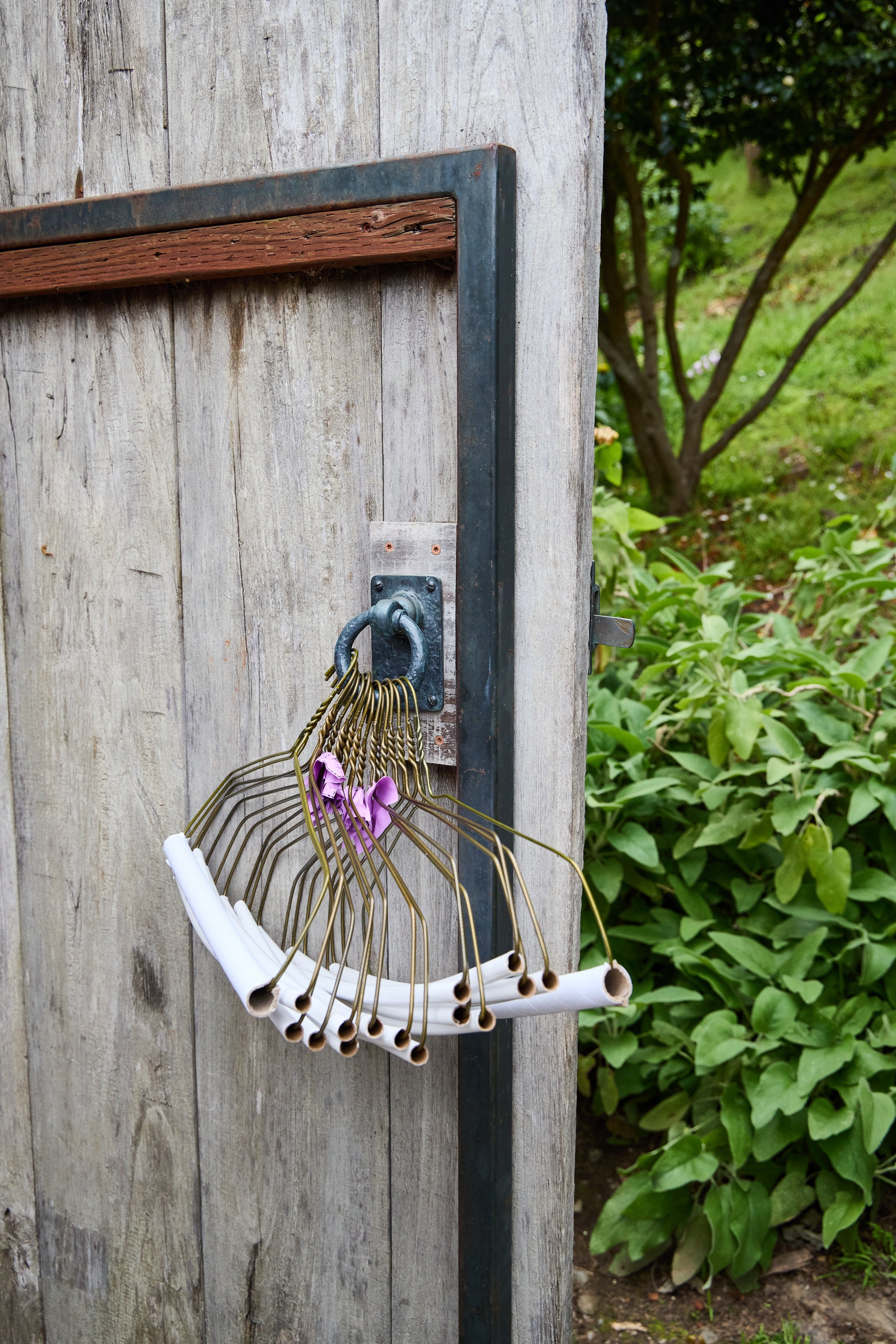 Close-up of a wooden outdoor post with a metal hook holding multiple wire hangers, some with rolled-up paper or cloth, situated next to greenery and bushes.