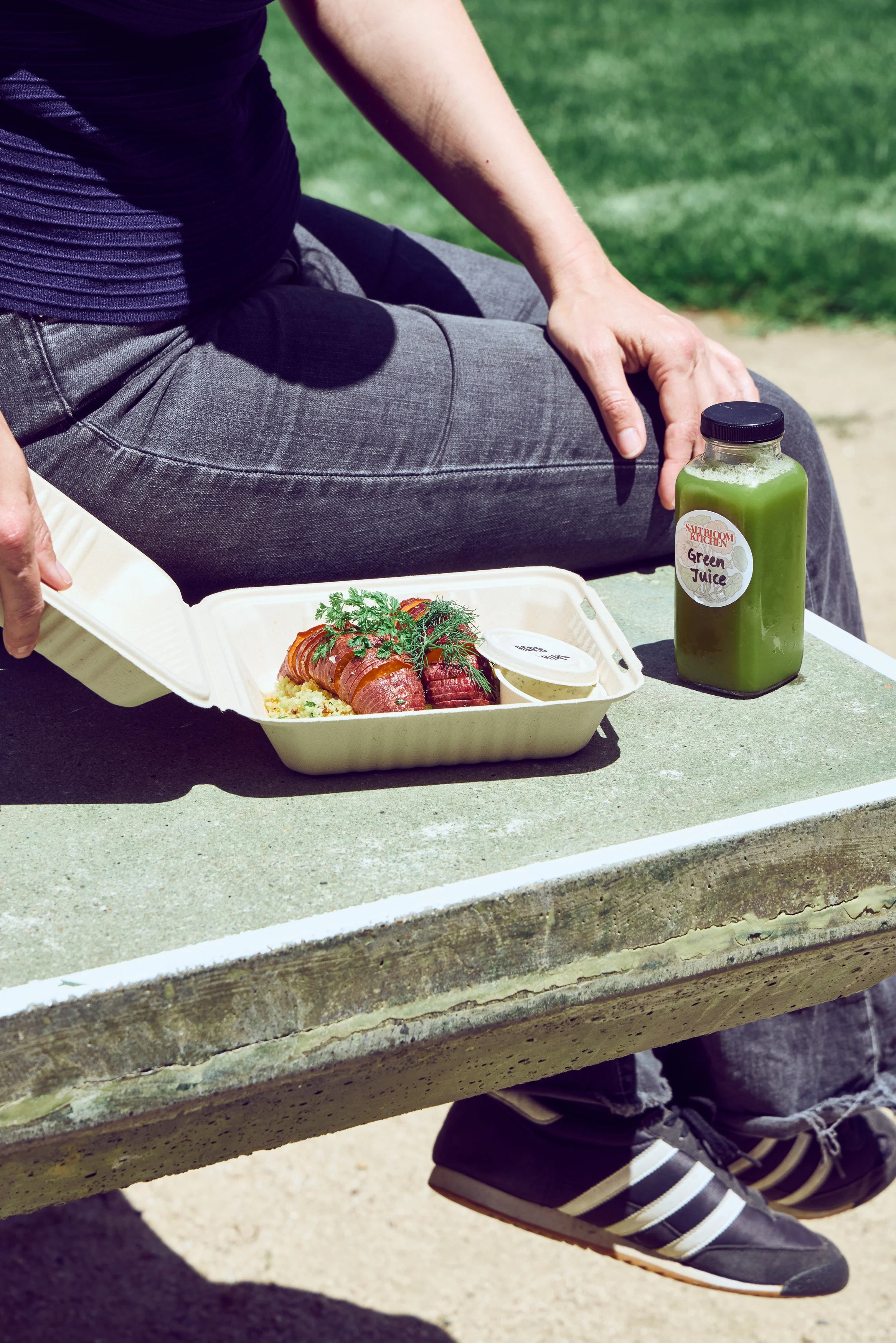 A person sitting on a park bench holding a takeout container with food and a bottle of green juice labeled "Saltbloom Kitchens Green Juice" from Salt Bloom Kitchen in Stinson Beach.