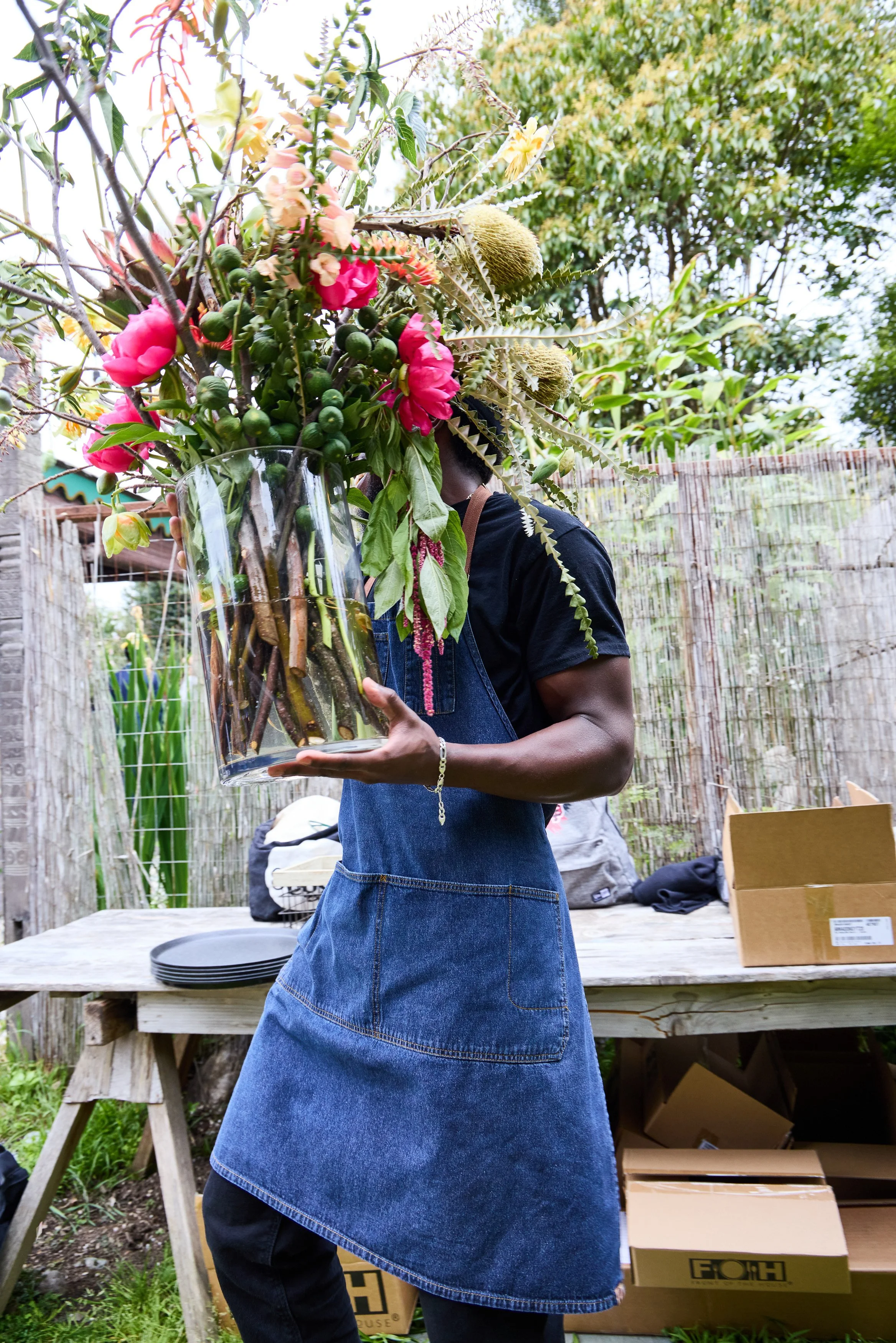 A person holding a large glass vase filled with an elaborate bouquet of various flowers, greenery, and branches, standing outdoors near a wooden table and garden fencing at Debonair Events wedding reception.