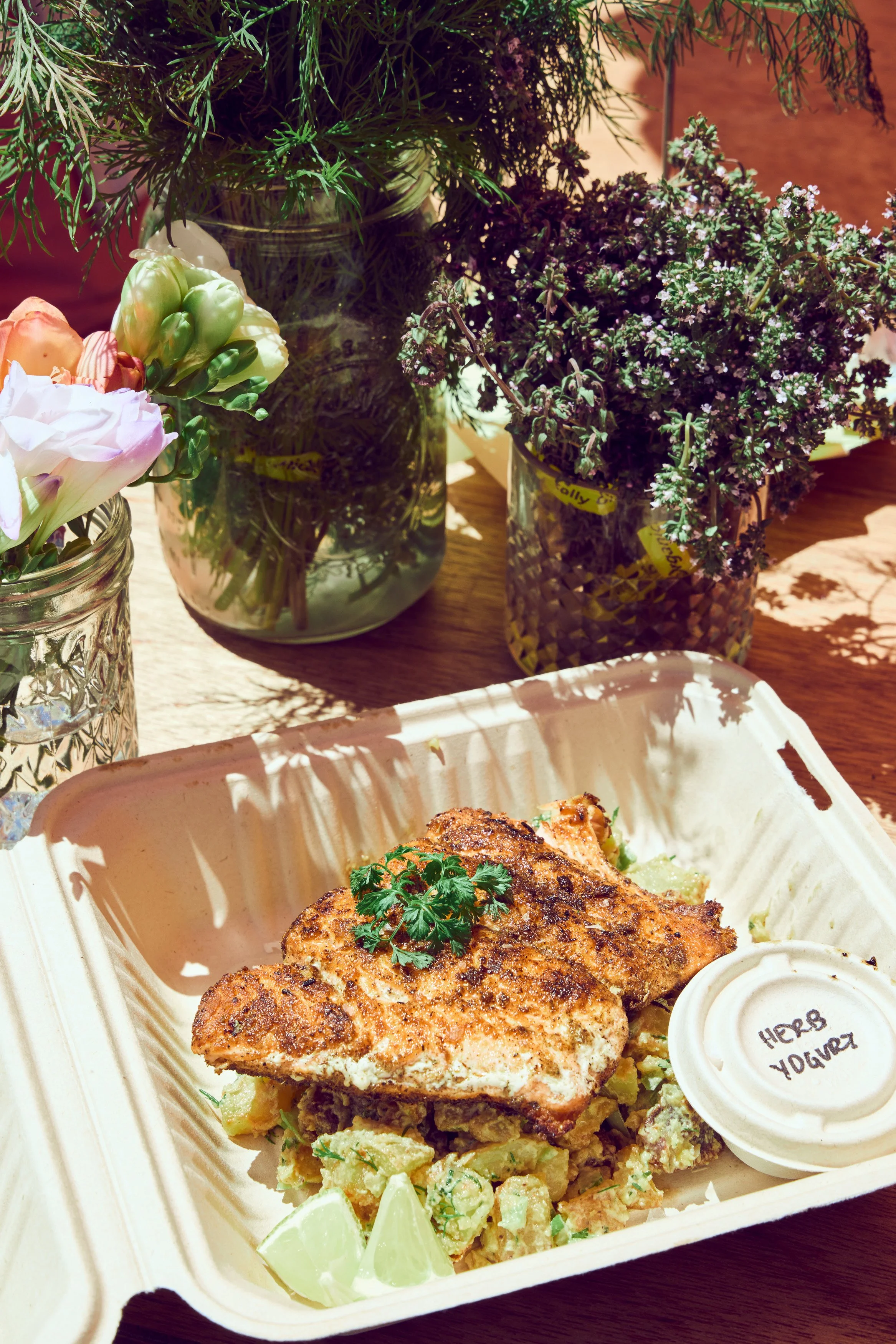 A Salt Bloom Cafe takeout box with a grilled salmon fillet garnished with cilantro, sliced lime, and served on a bed of summer potato salad with herbs. In the background, there are jars with assorted flowers and greenery.