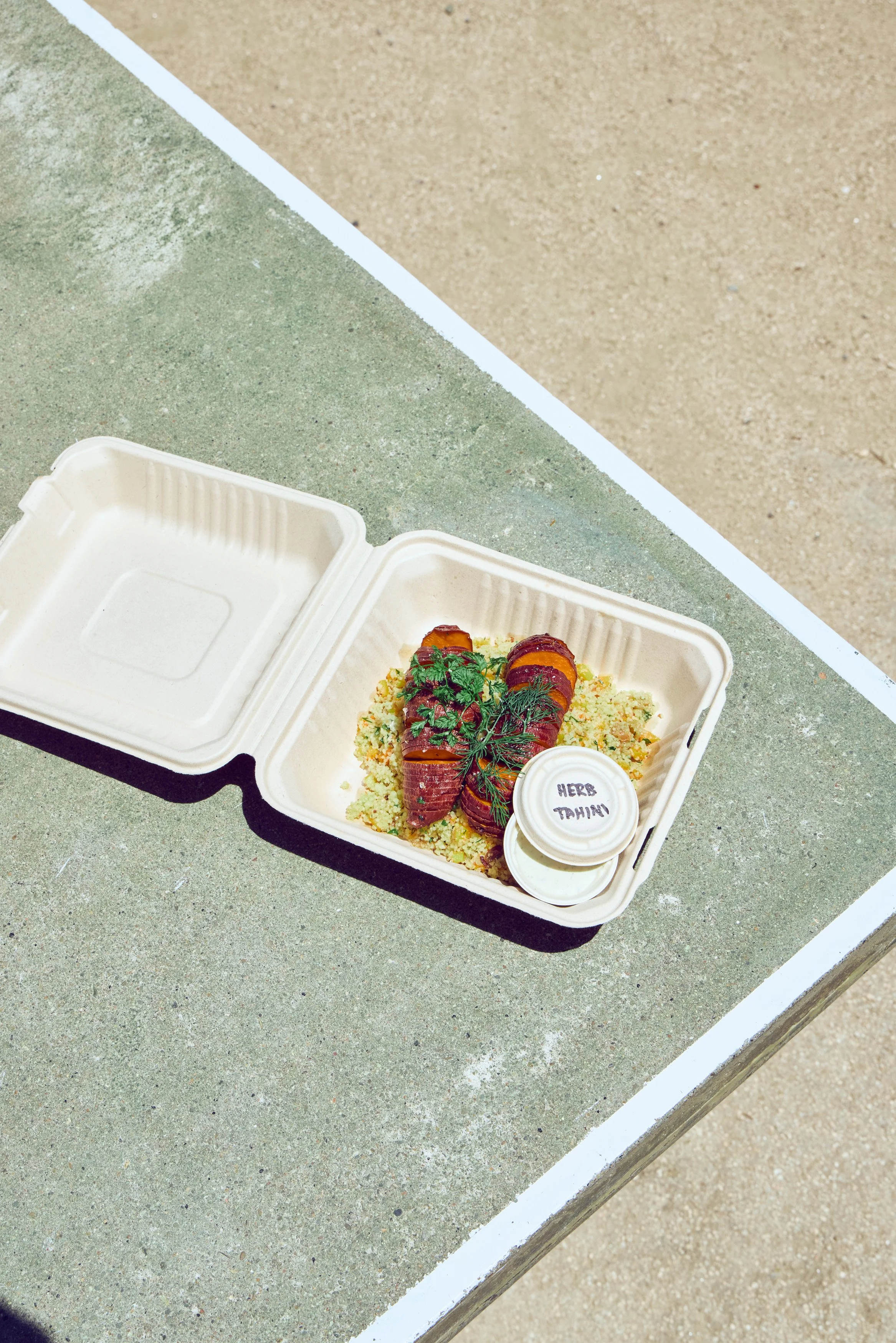 Open takeout container with baked Salt Bloom Kitchen sweet potatoes topped with fresh herbs, served over a bed of couscous on a beachside table.