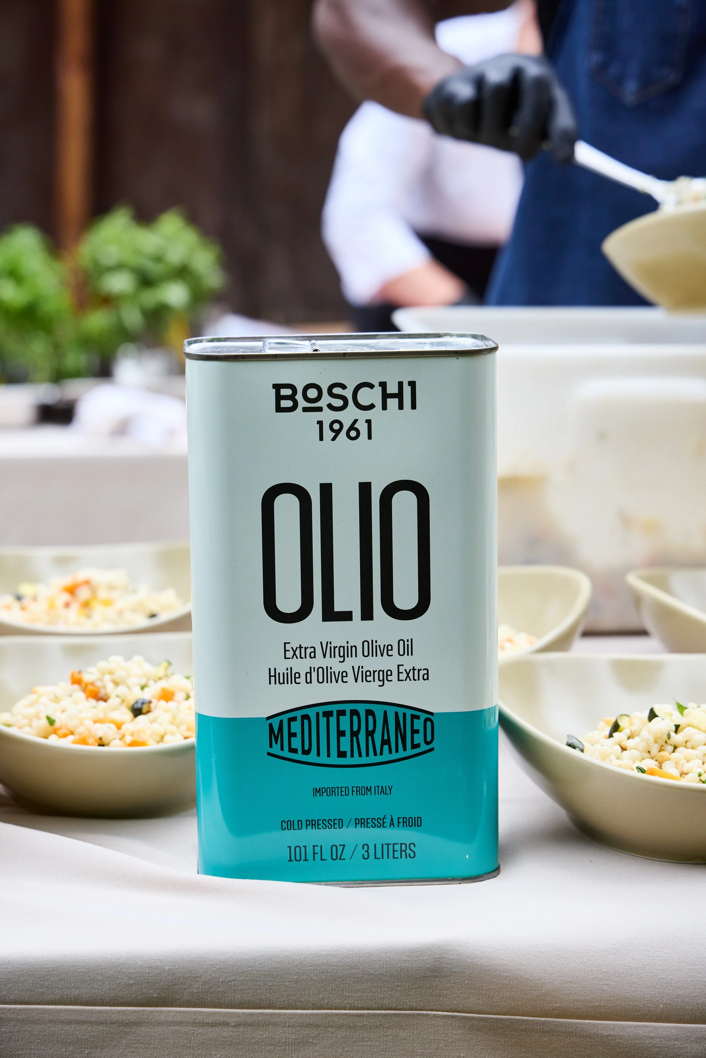 A can of Boschi extra virgin olive oil with a light blue and white label in front of bowls of cous-cous on a table, with a blurry Debonair Events chef in the background.