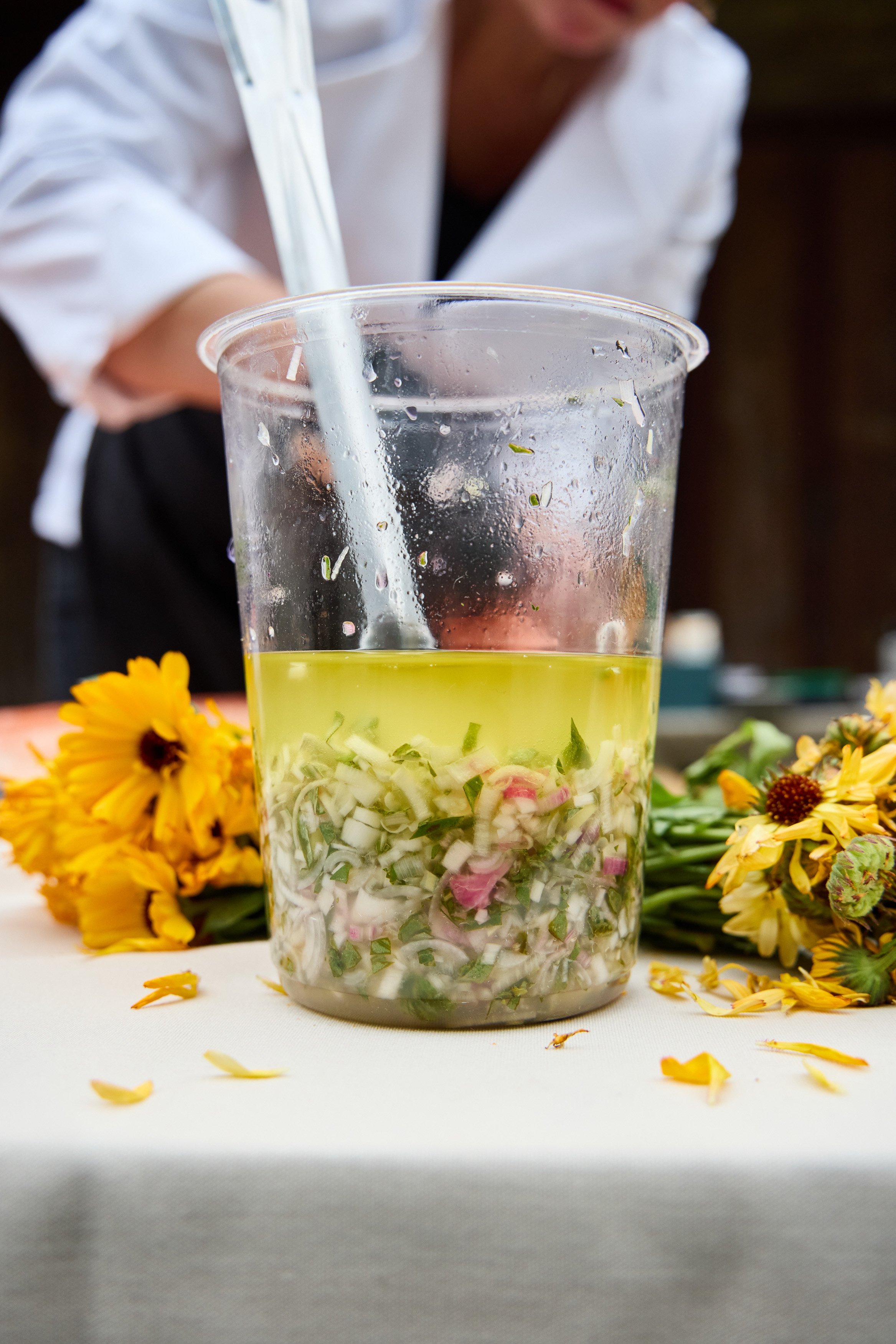Close-up of a catering prep container filled with chopped vegetables and olive oil, with yellow flowers scattered around on a white surface, and Debonair Events Chef in a white shirt in the background.