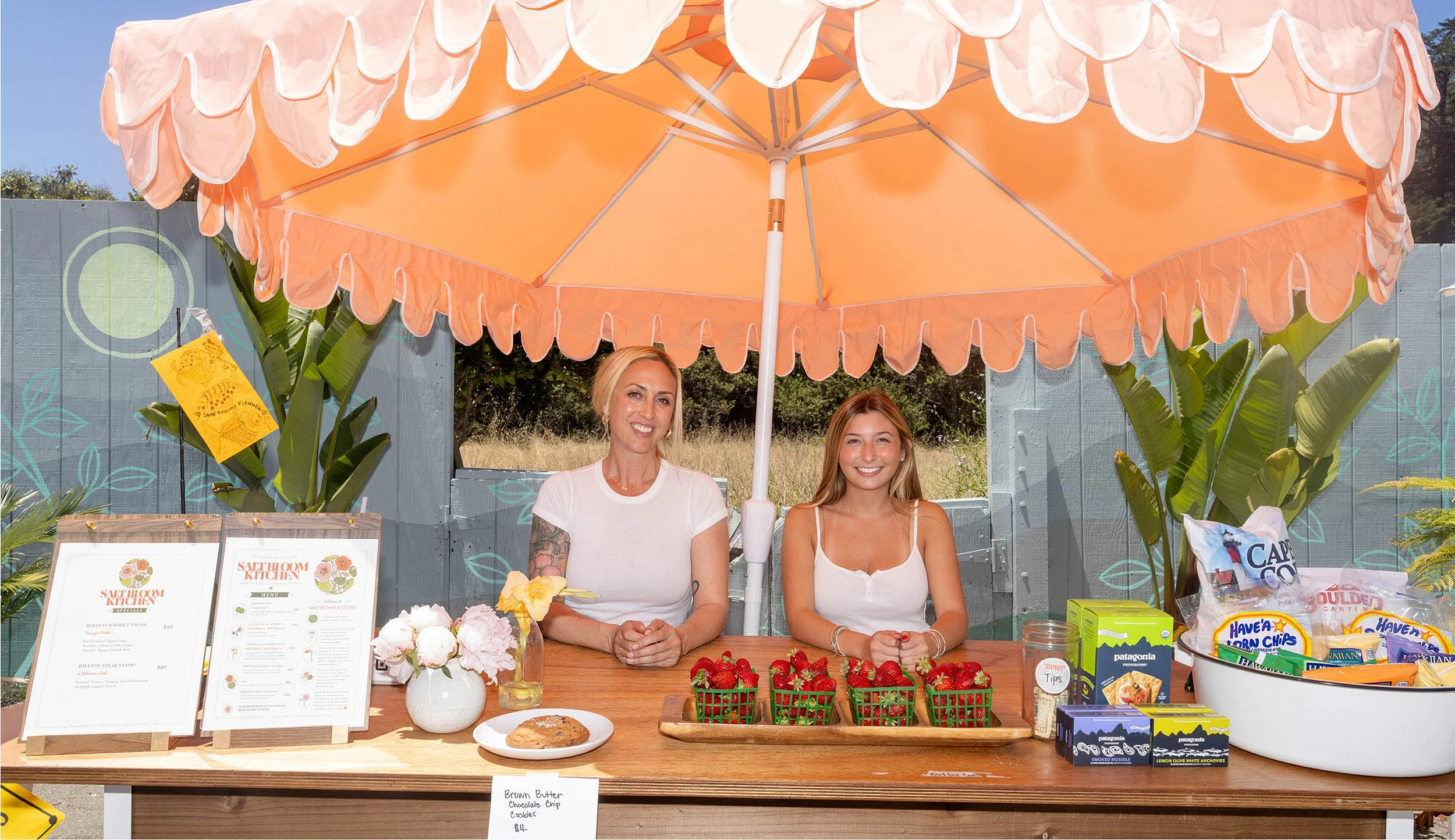 Debonair founder Rachel DiPaolo and daughter stand under peach colored umbrella at Salt Bloom Cafe summer pop-up store front.