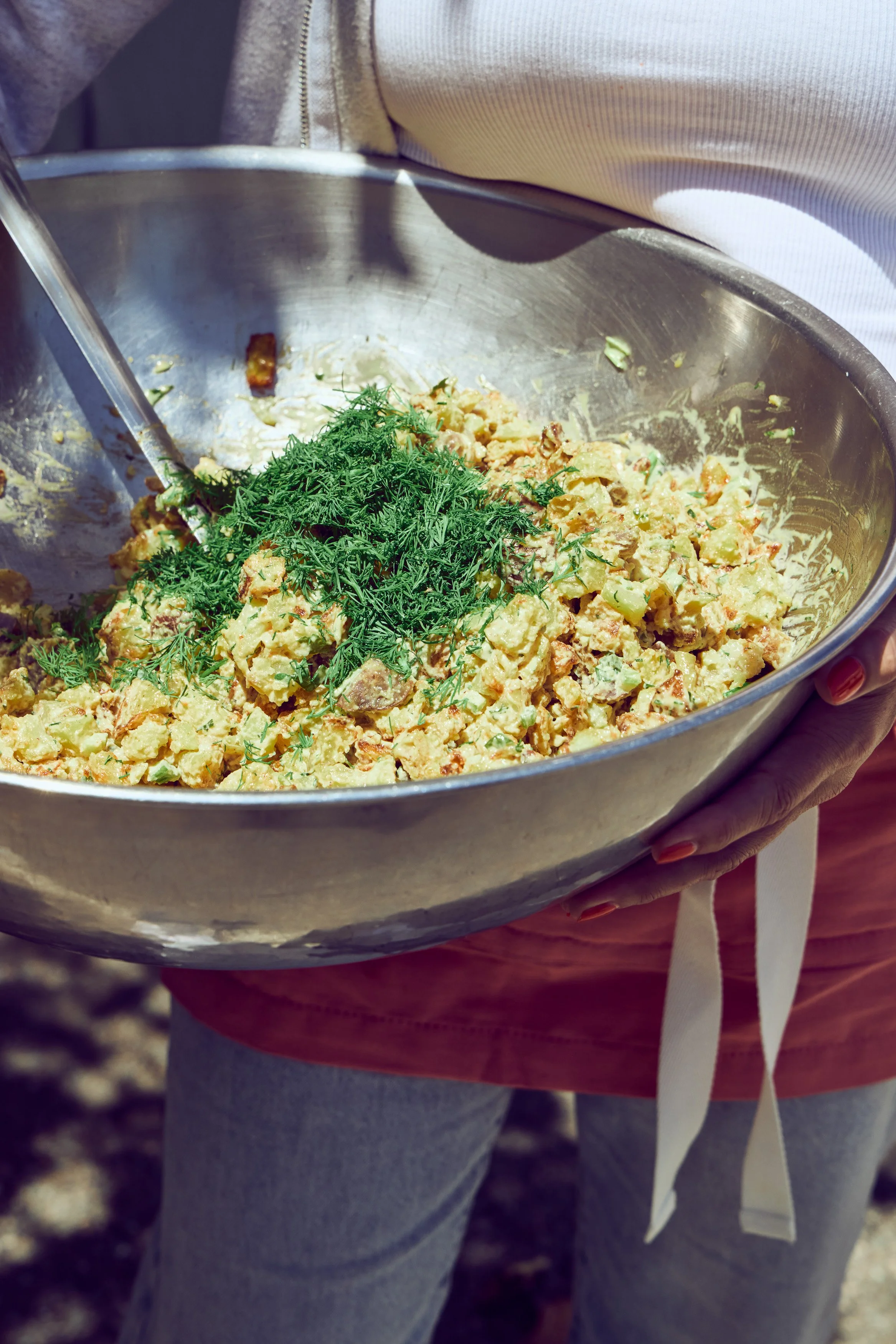Rachel DiPaolo holding a large metal bowl filled with summer potato salad, mixing in fresh herbs.