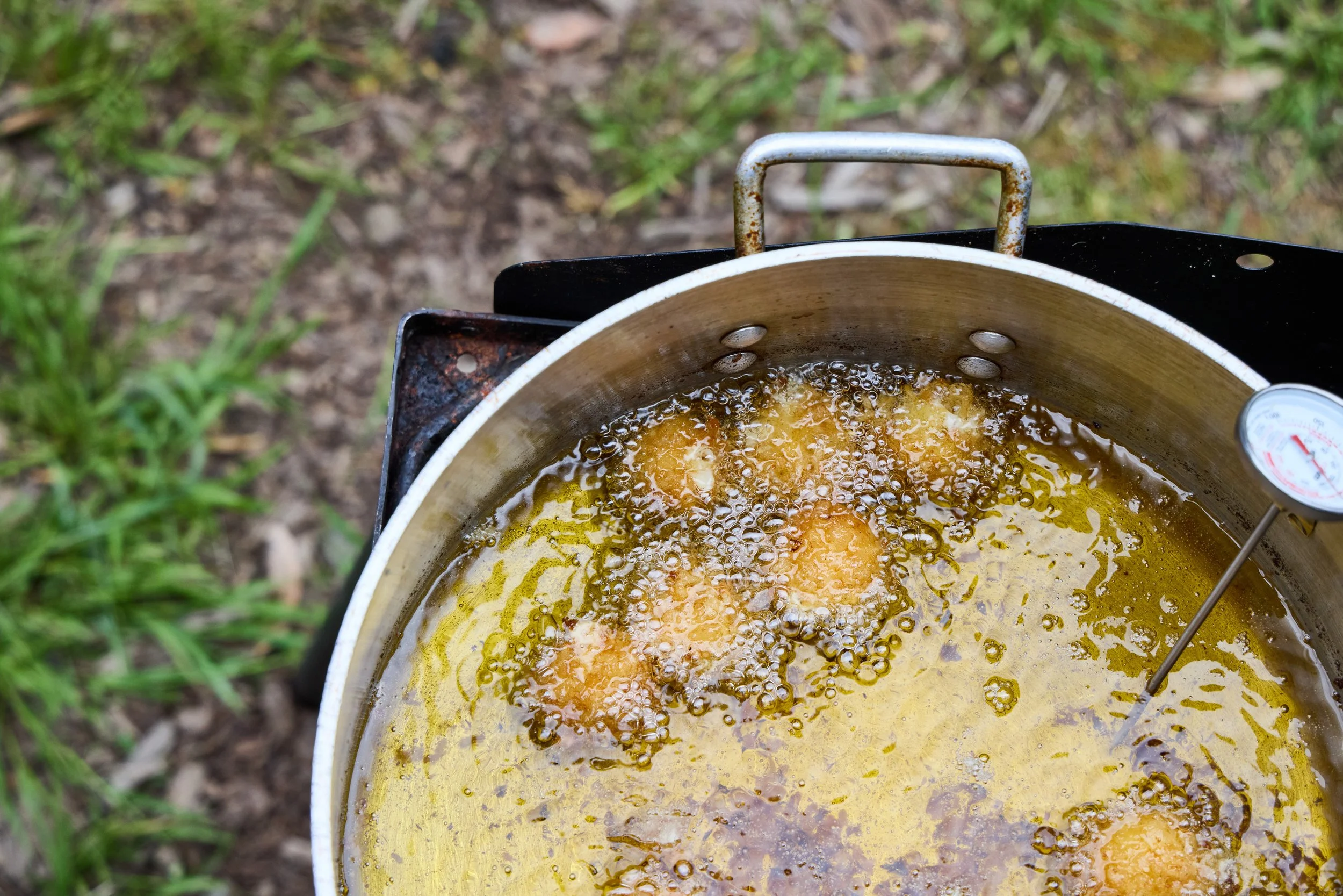 A large, round frying pan on a portable stove outdoors, with bubbling hot oil and fritters frying inside.