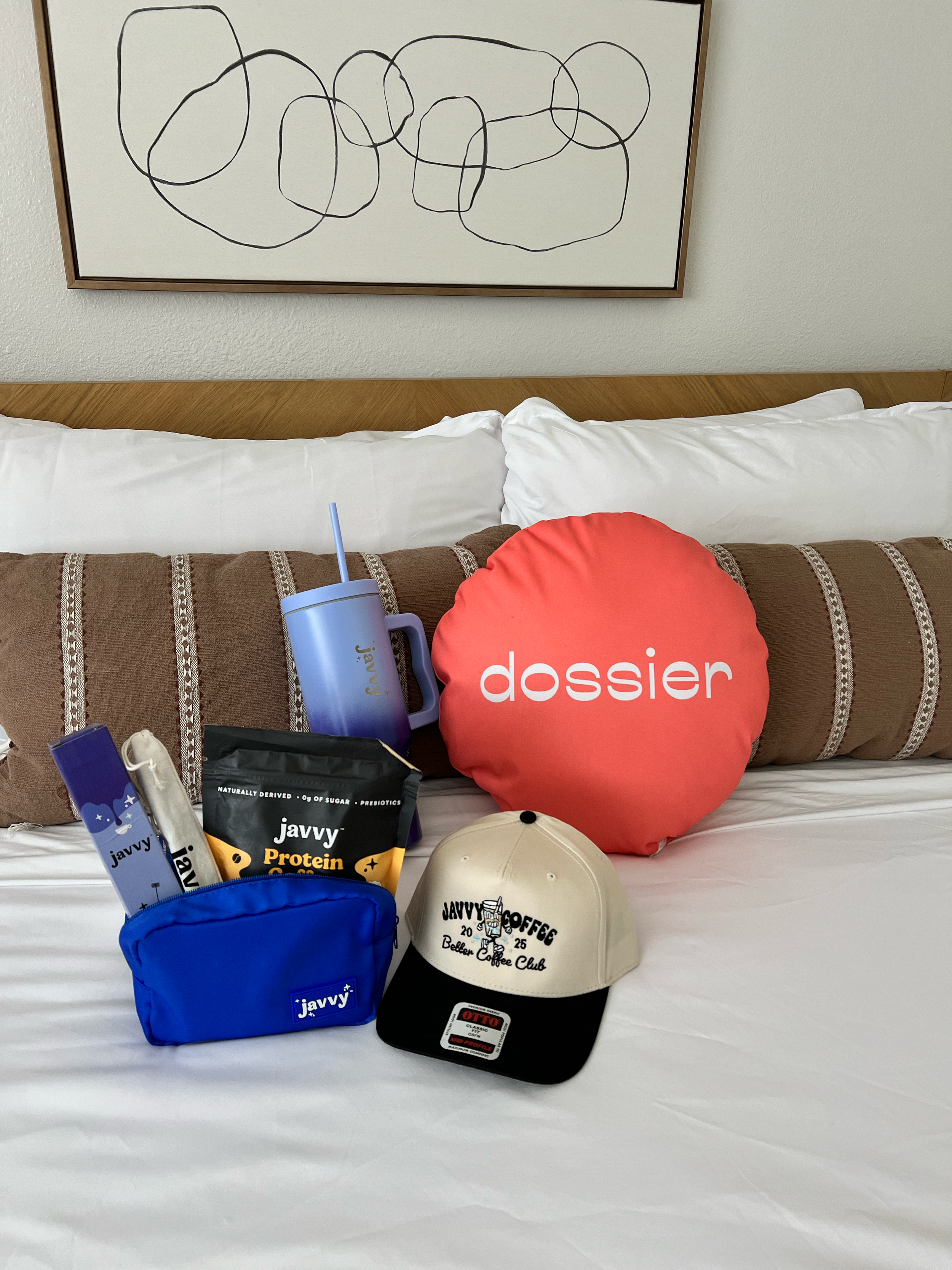 A hotel bed with a white sheet, various items including a blue pouch labeled 'javy', a black hat with 'Javy Coffee' logo, a beige cap, a red puff cushion with 'dossier' text, a blue stainless steel tumbler, and some snack packets. There is a framed a