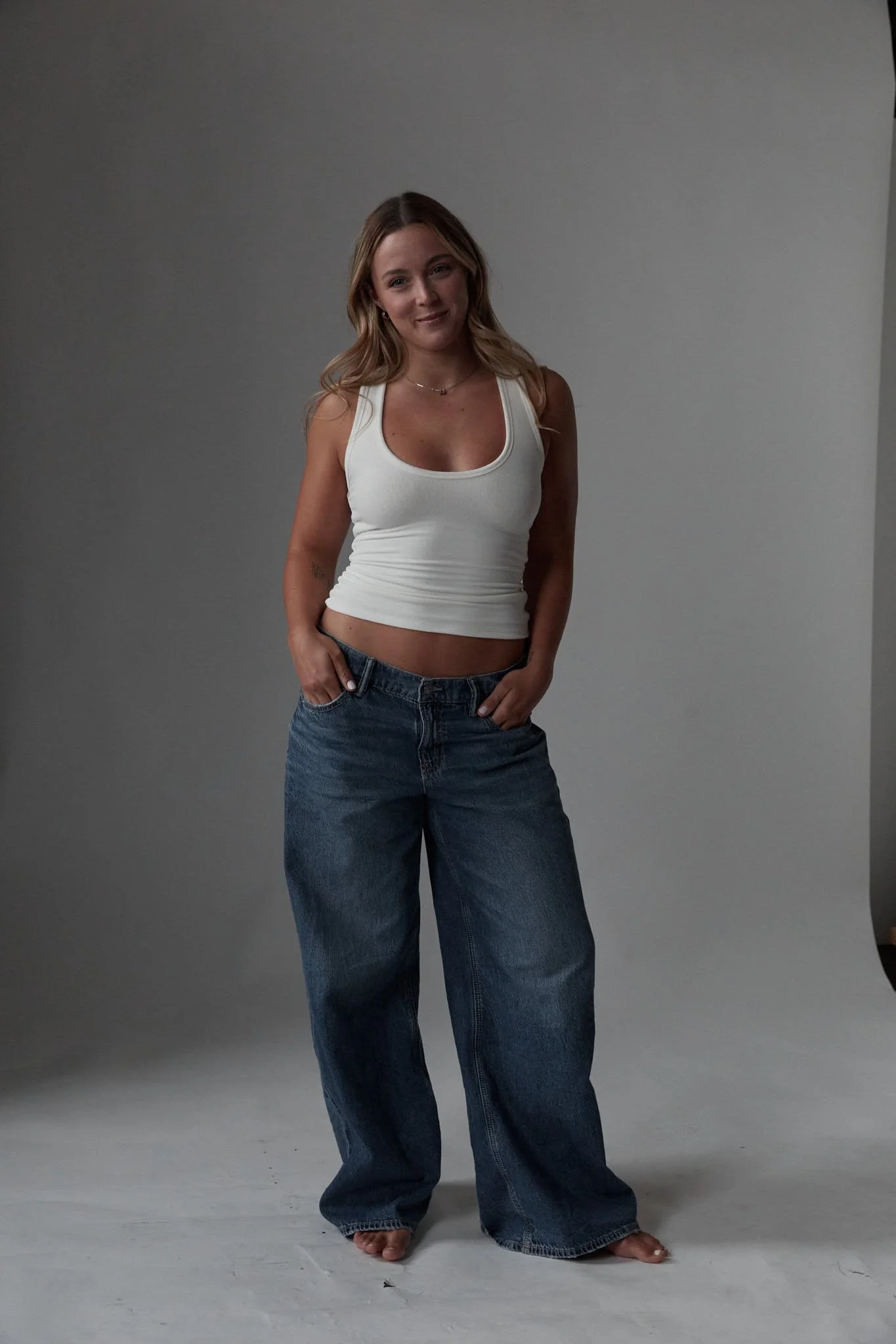 A woman with long light brown hair wearing a white tank top and high-waisted blue jeans, barefoot, stands in front of a plain gray background, smiling gently.