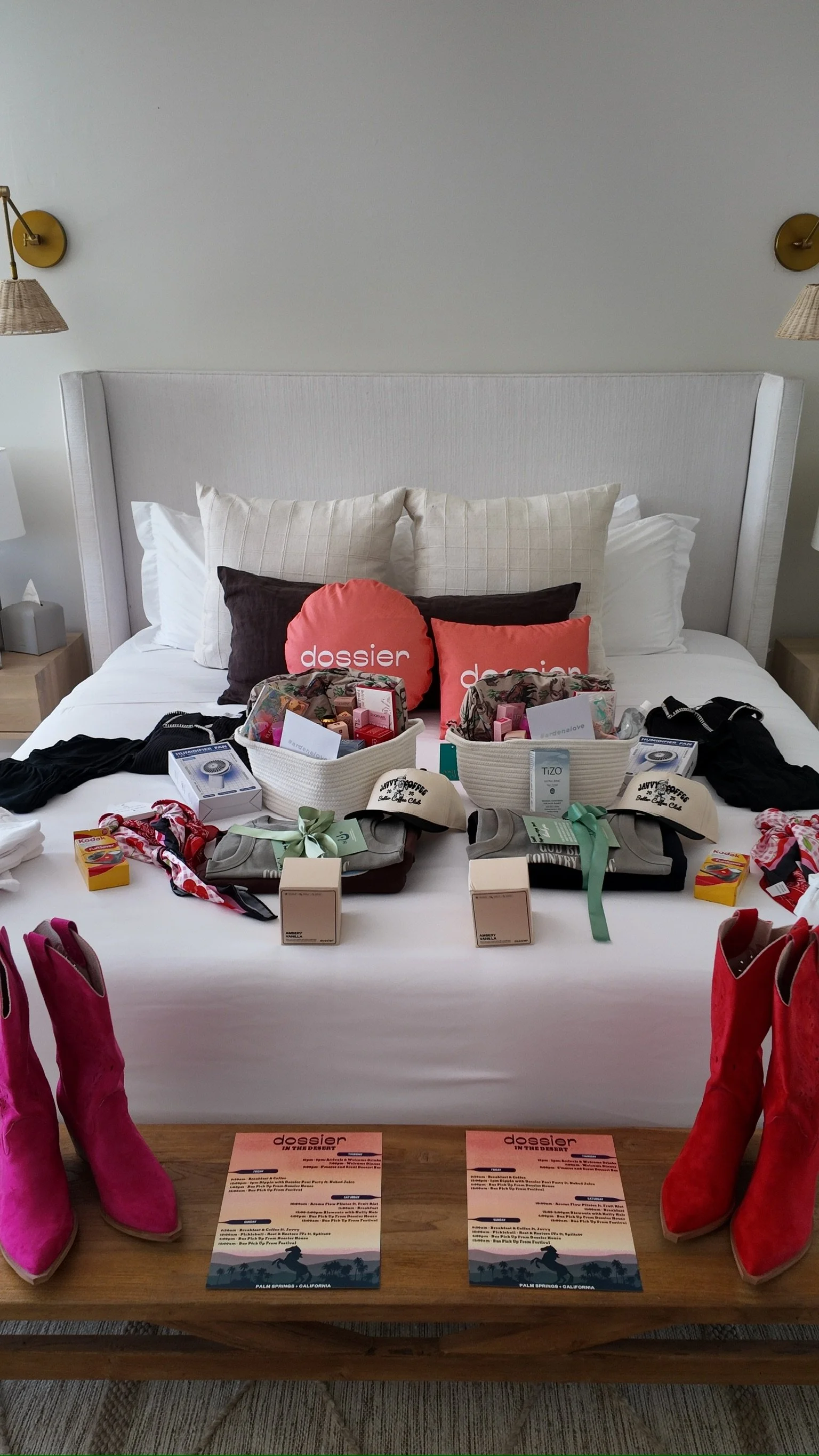 A bed with various items on it, including shoes, boxes, and toiletry items, in a hotel room with pillows and wall lamps.