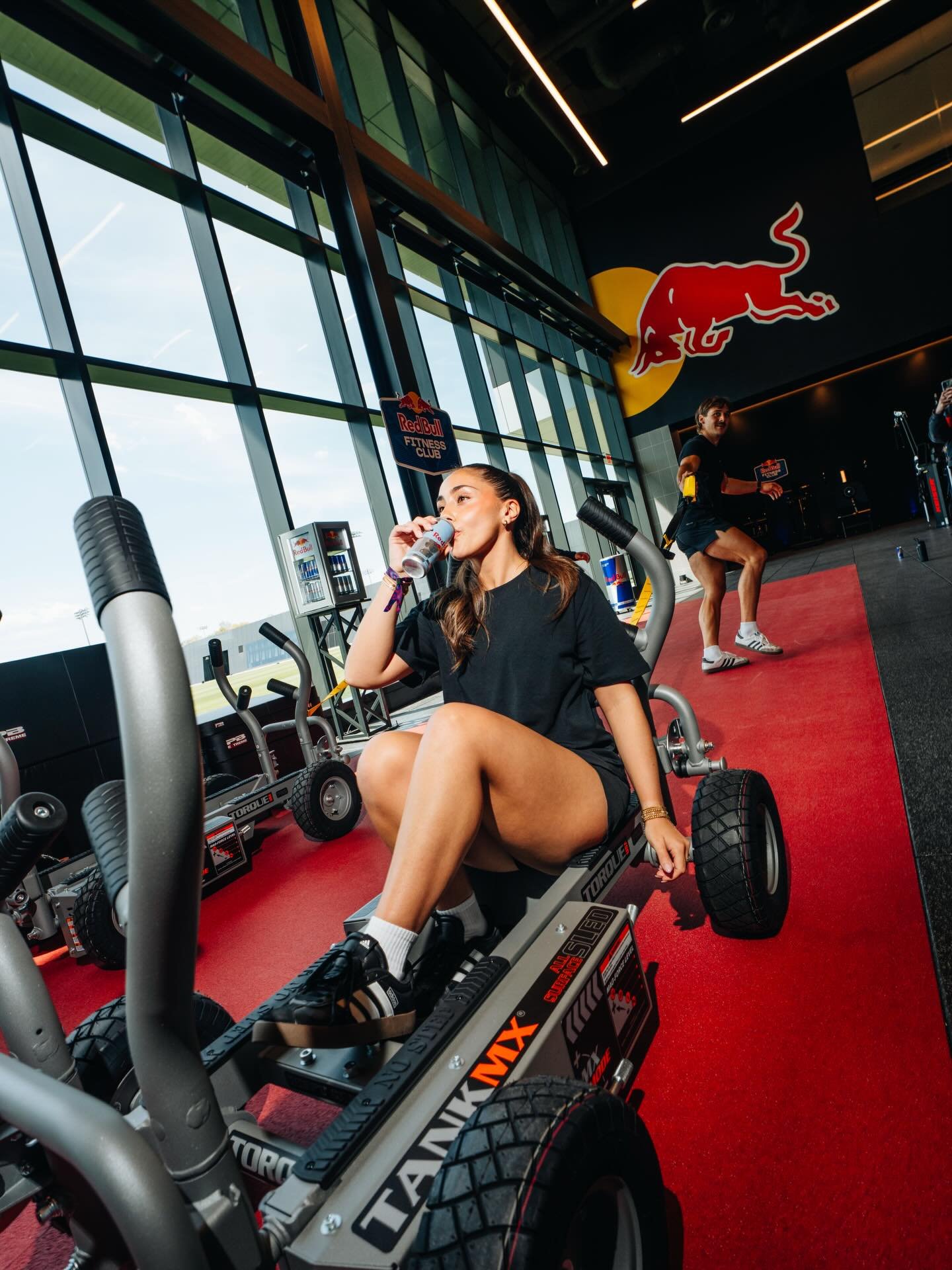 A woman sitting on an exercise bike drinking from a Red Bull energy drink in a gym with large windows and a Red Bull logo on the wall, and another woman in the background doing exercises.