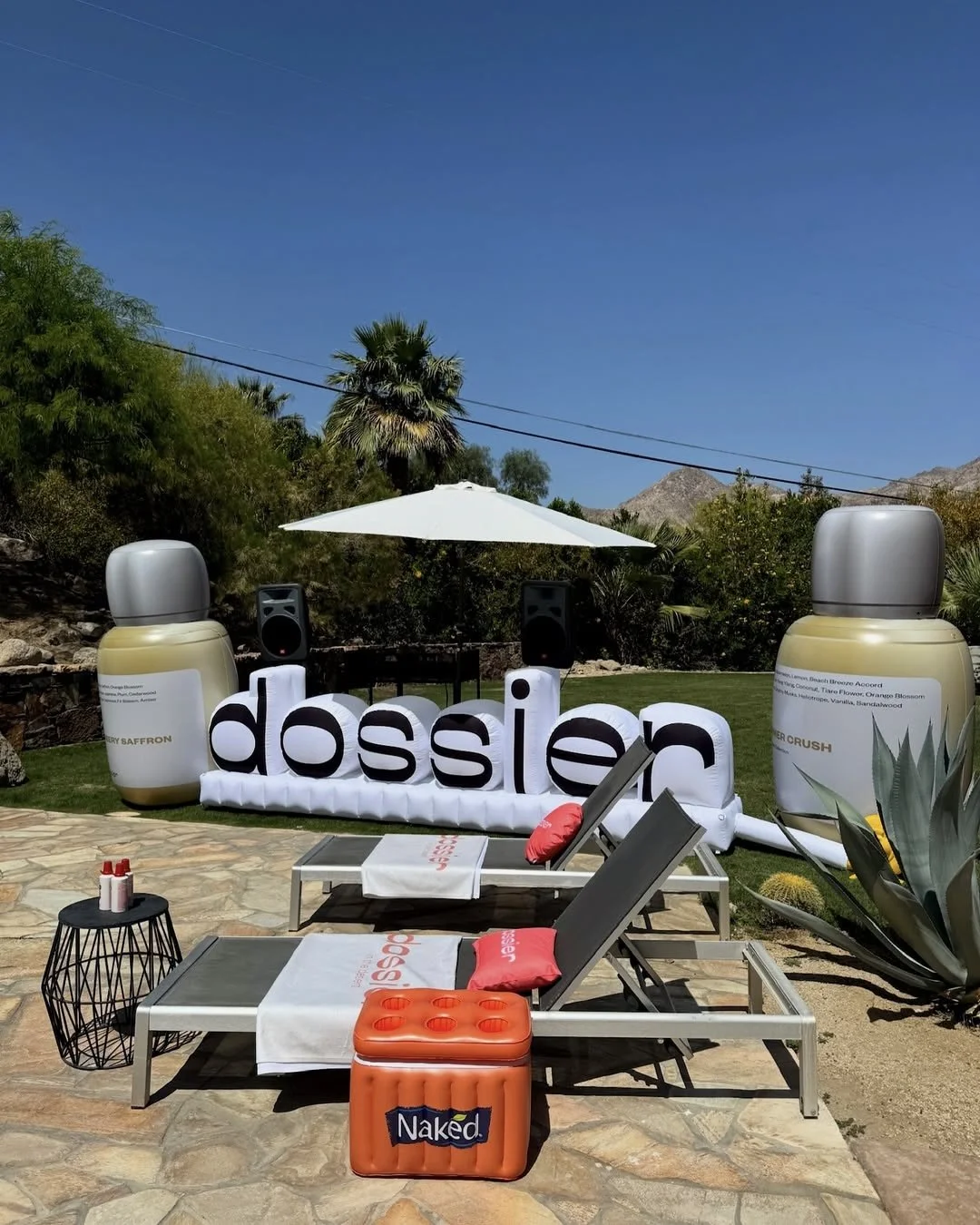 Outdoor poolside setting with lounge chairs, a white umbrella, large decorative bottle props, a sign that reads 'dossier', and an orange cooler labeled 'Naked' against a backdrop of trees, mountains, and a blue sky.