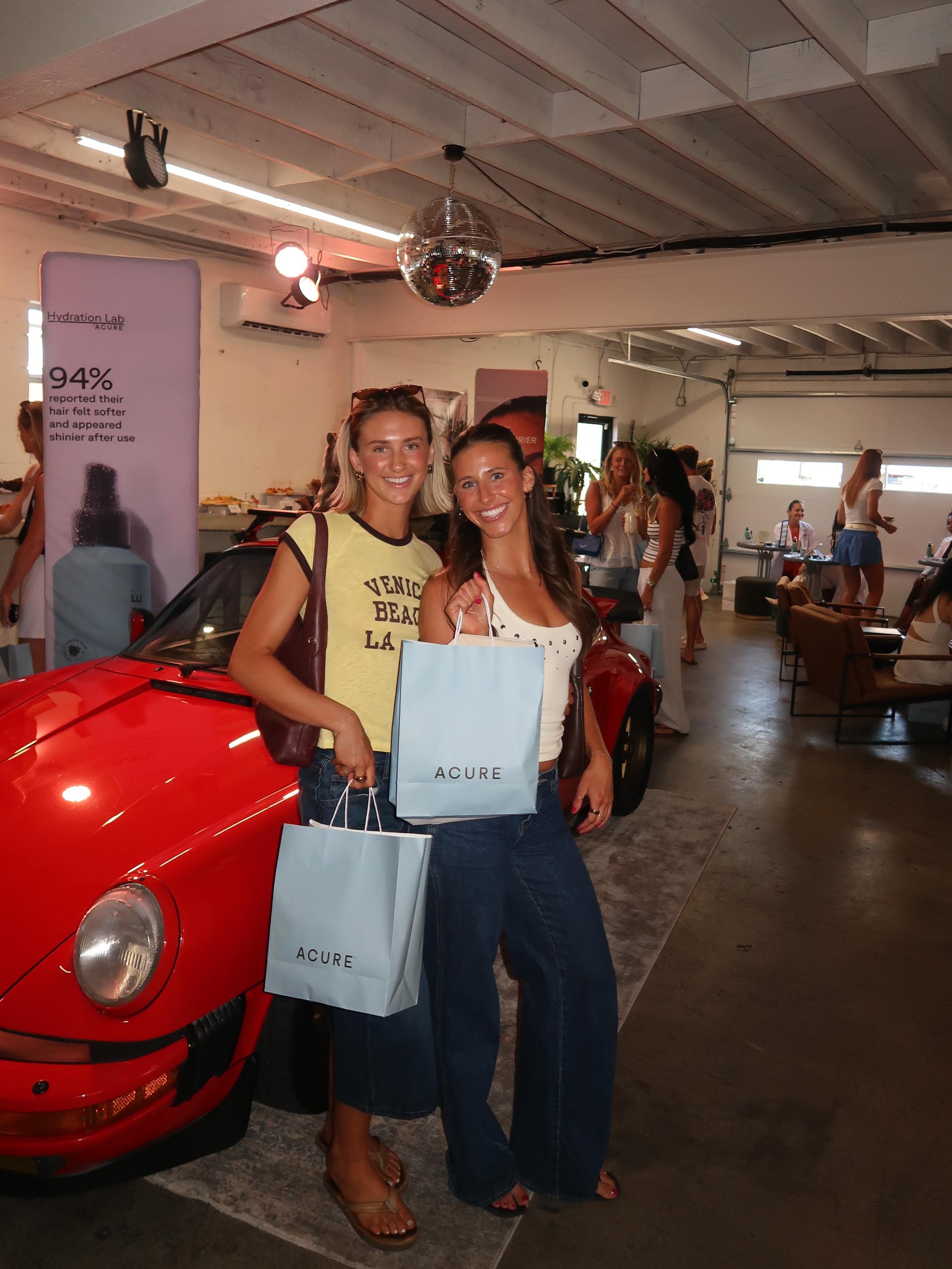 Two smiling women standing in front of a red sports car inside a modern industrial style building, holding shopping bags with the ACURE logo, with other people socializing in the background.
