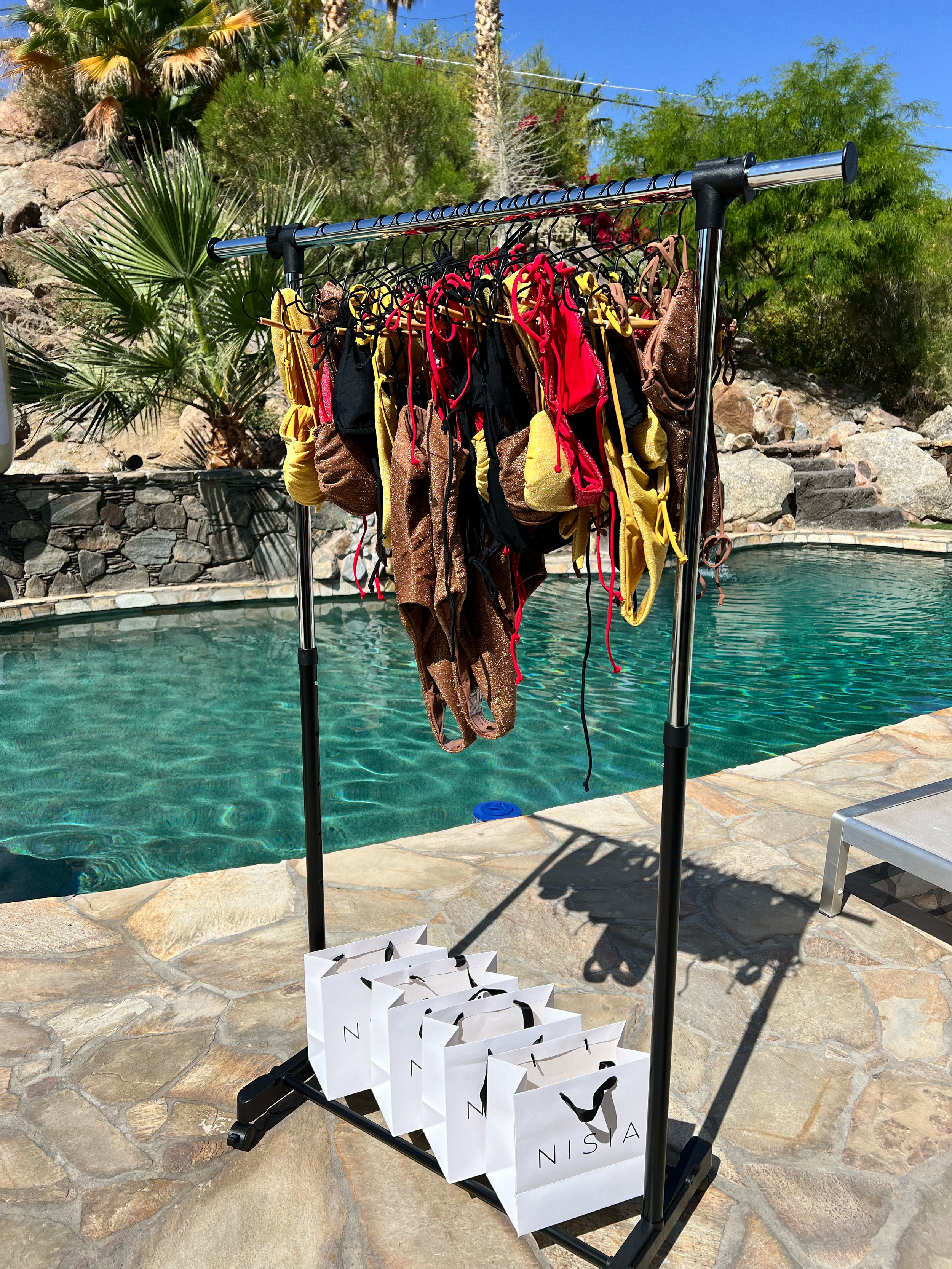 A clothing rack with various swimsuits hanging on it, positioned outside by a pool with a stone patio, surrounded by desert plants and rocks, sunny weather, white shopping bags with "NISSA" written on them placed under the rack.