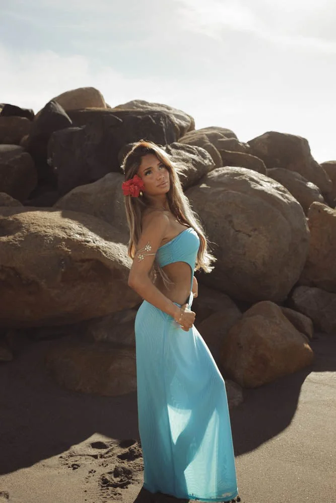 A woman in a blue strapless dress standing on a sandy beach with large rocks in the background, wearing a red flower in her hair.