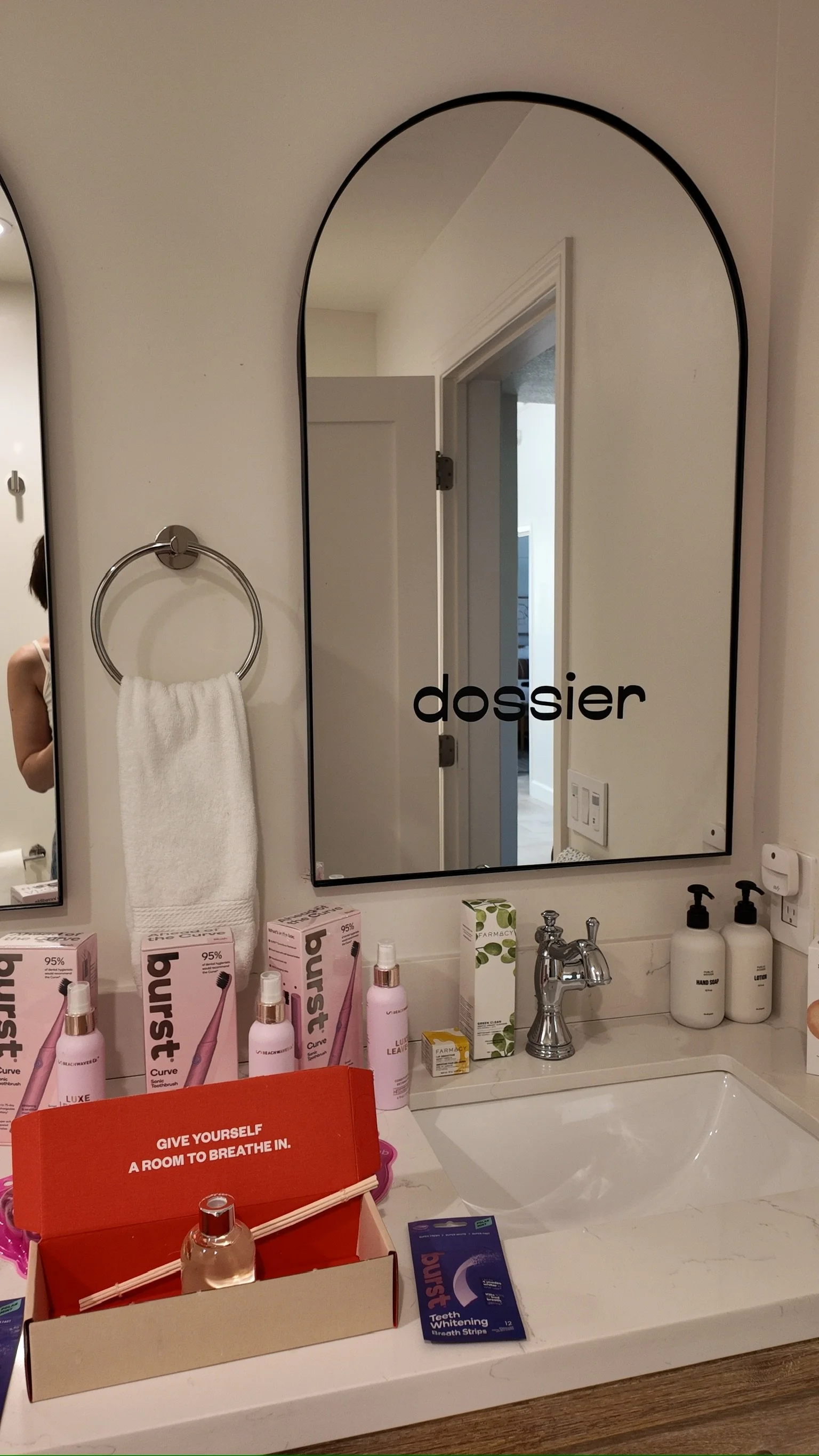 Bathroom bathroom mirror reflection showing a doorway, wall switch, and part of a person taking the photo. Items on the bathroom counter include pink electric toothbrushes, a red brochure, hygiene products, and a box of teeth whitening strips.