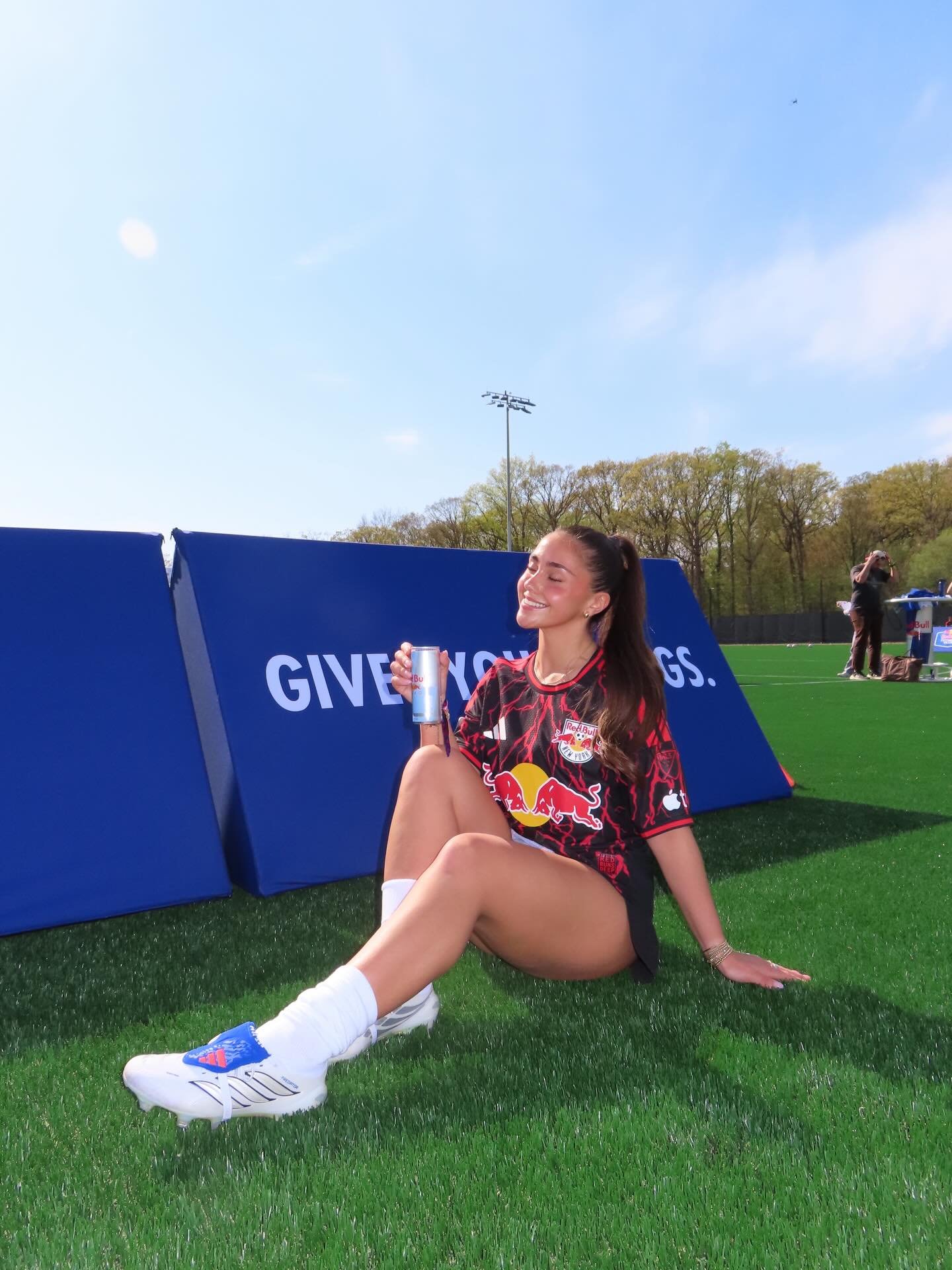A young woman in a Red Bull soccer jersey is sitting on green turf with her eyes closed and smiling while holding a drink. She is wearing white soccer cleats and has long dark hair tied back. Behind her, there is a blue barrier with white text partially visible that reads "GIVE YOUR G..." and a few people are in the background, including a person taking photos.