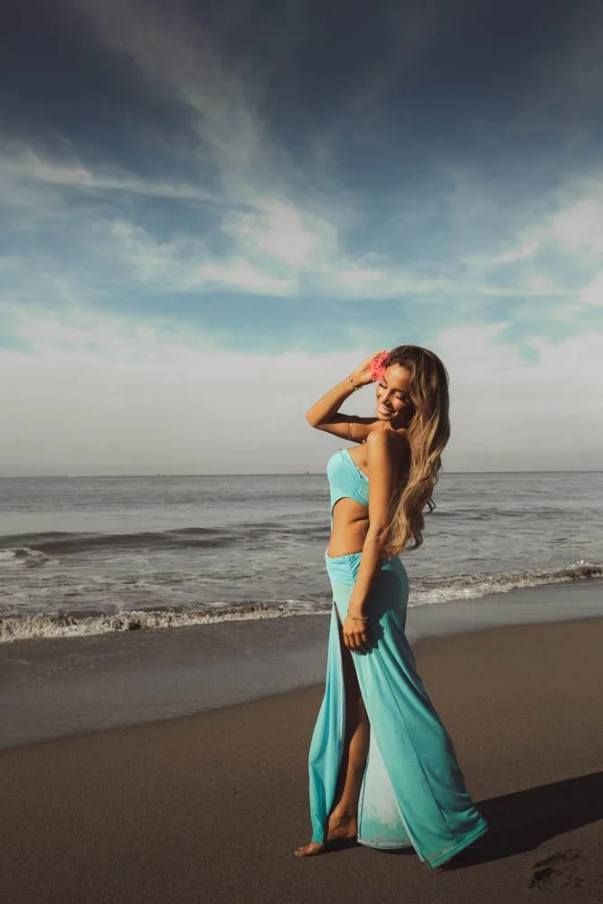 Young woman smiling on the beach wearing a turquoise dress, holding a seashell to her ear, with the ocean and sky in the background.