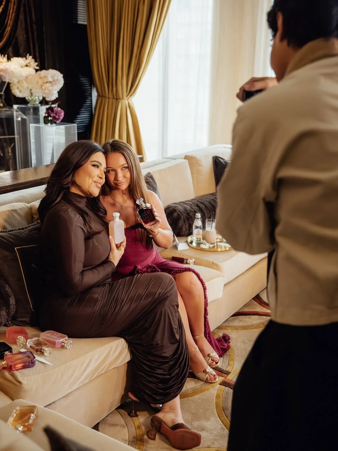 Two women sit on a beige sofa in a luxurious room, holding perfume bottles and smiling at a photographer taking their photo. The woman on the left wears a dark brown dress, and the woman on the right wears a maroon dress. Various perfume bottles are on the sofa and nearby tables, with a window and curtains in the background.