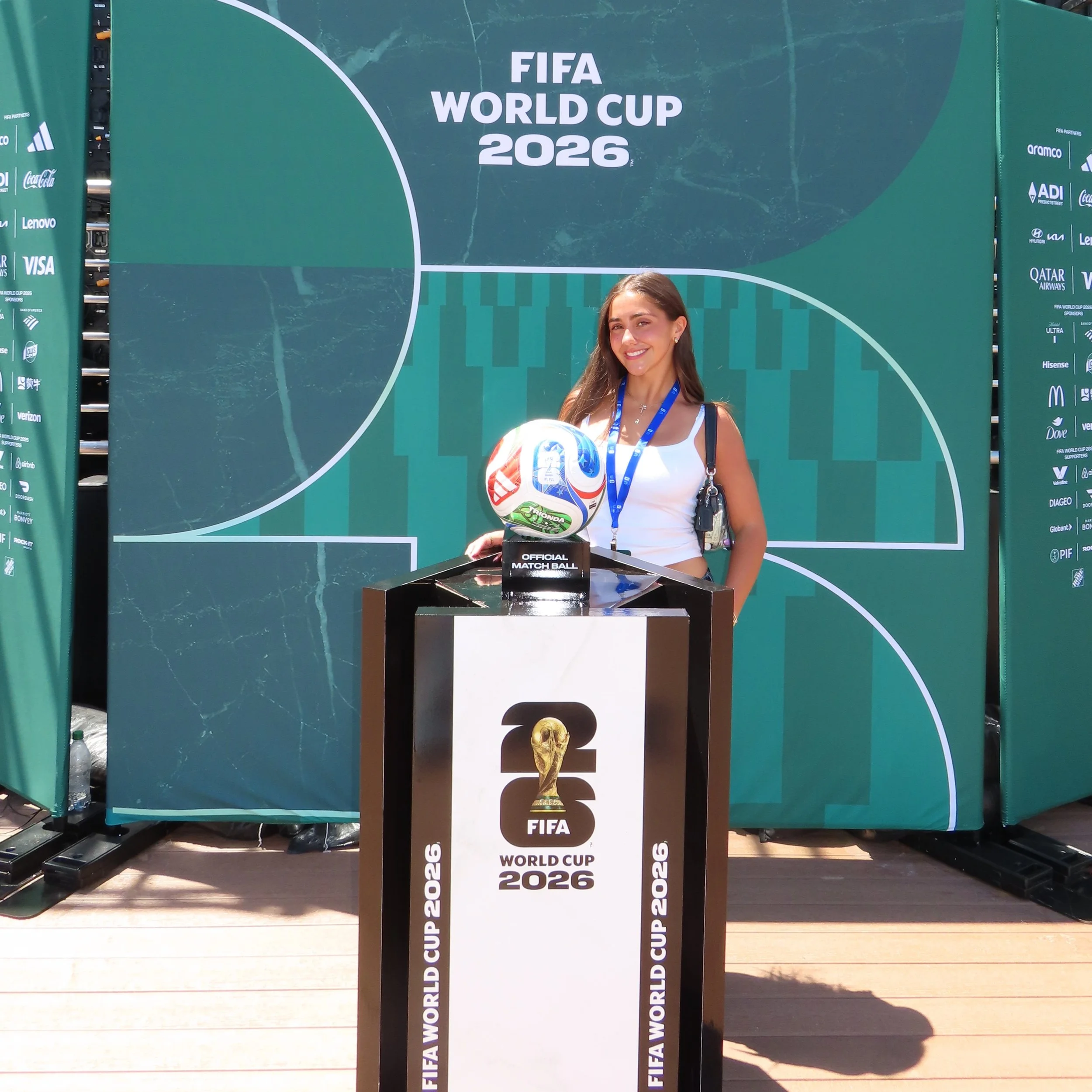 A woman standing next to a display with the FIFA World Cup 2026 logo and an official match ball. She is smiling, wearing a white top, and has a lanyard around her neck. The background shows a large green FIFA World Cup 2026 promotional banner and various sponsor logos.