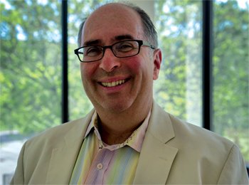 A smiling man with glasses wearing a light-colored suit and a pastel striped shirt, standing in front of a window with trees outside.