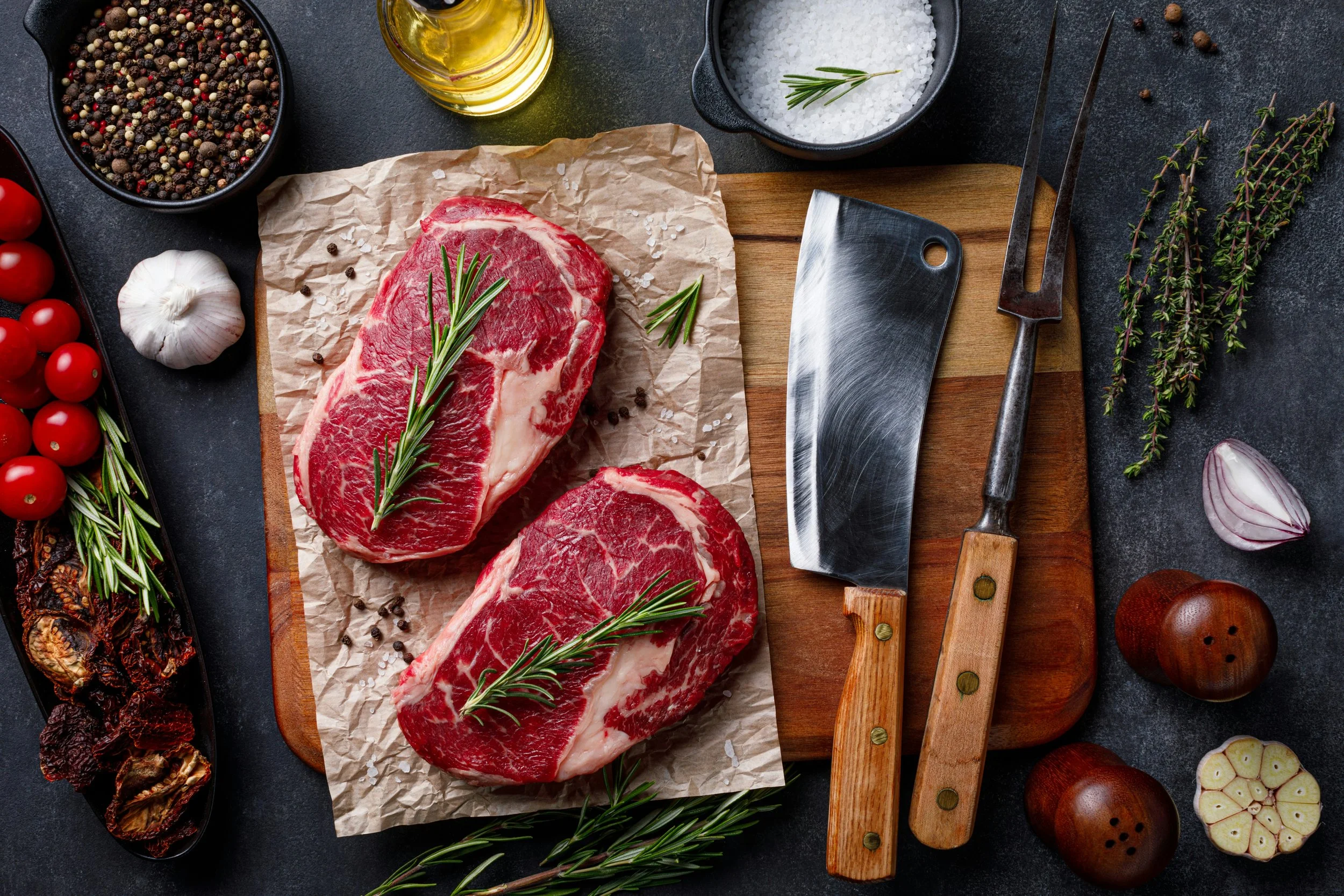 Raw marbled beef steaks on brown parchment paper on a chef's cutting board, surrounded by herbs, spices, garlic, cherry tomatoes, and kitchen tools on a dark surface.