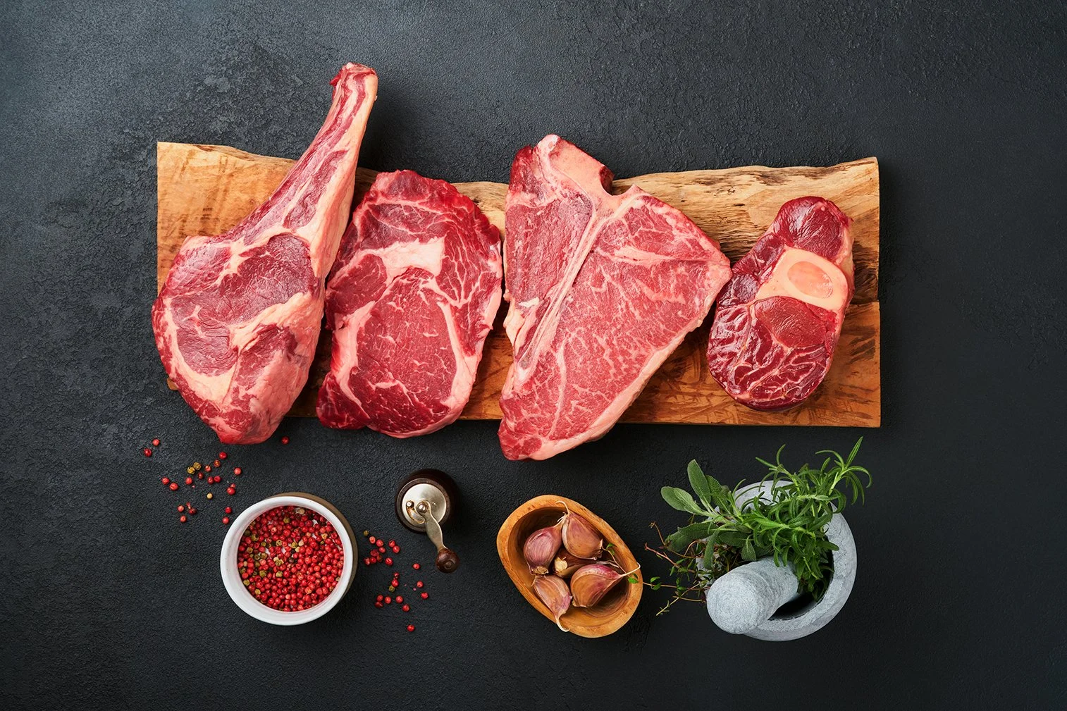 Various cuts of raw beef on a wooden board, surrounded by seasonings, garlic, and herbs on a black surface.