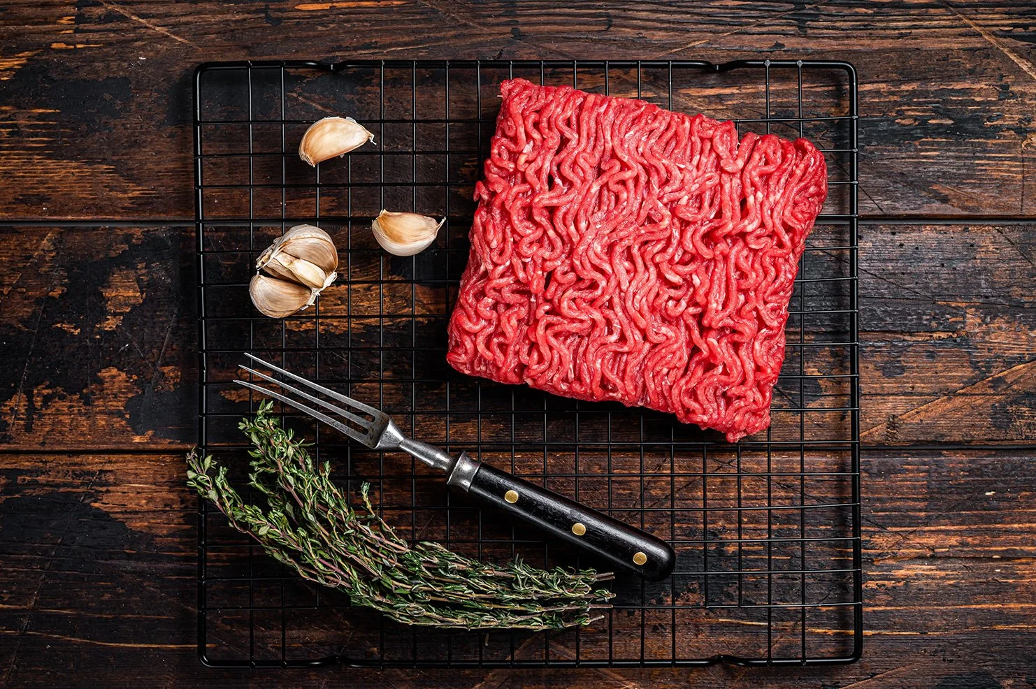 Raw ground beef, garlic cloves, a sprig of thyme, and a meat fork on a black wire rack on a wooden surface.