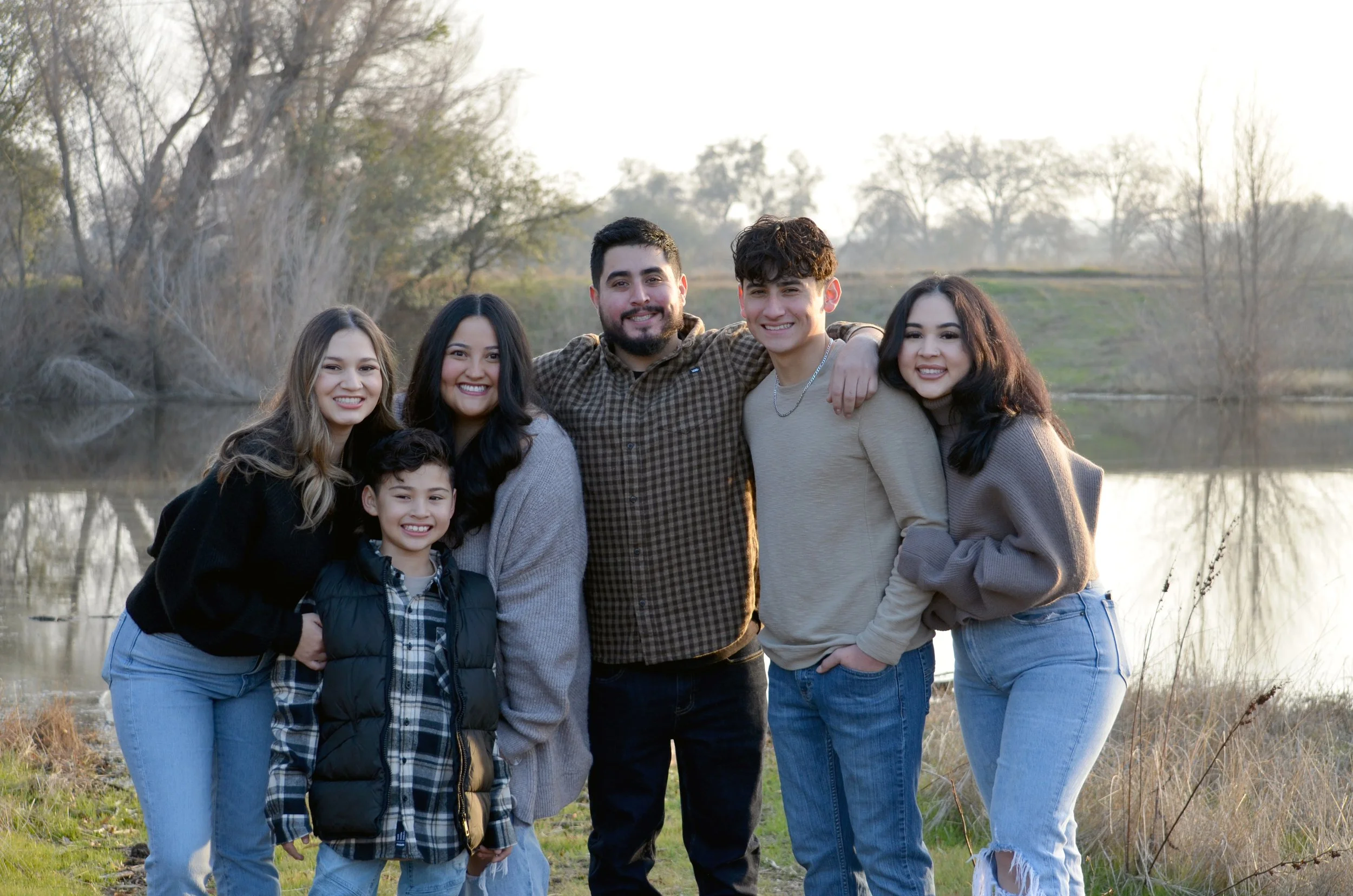 A group of six people, five women and one man, standing outdoors near a body of water with trees in the background at sunset. They are smiling and embracing each other.