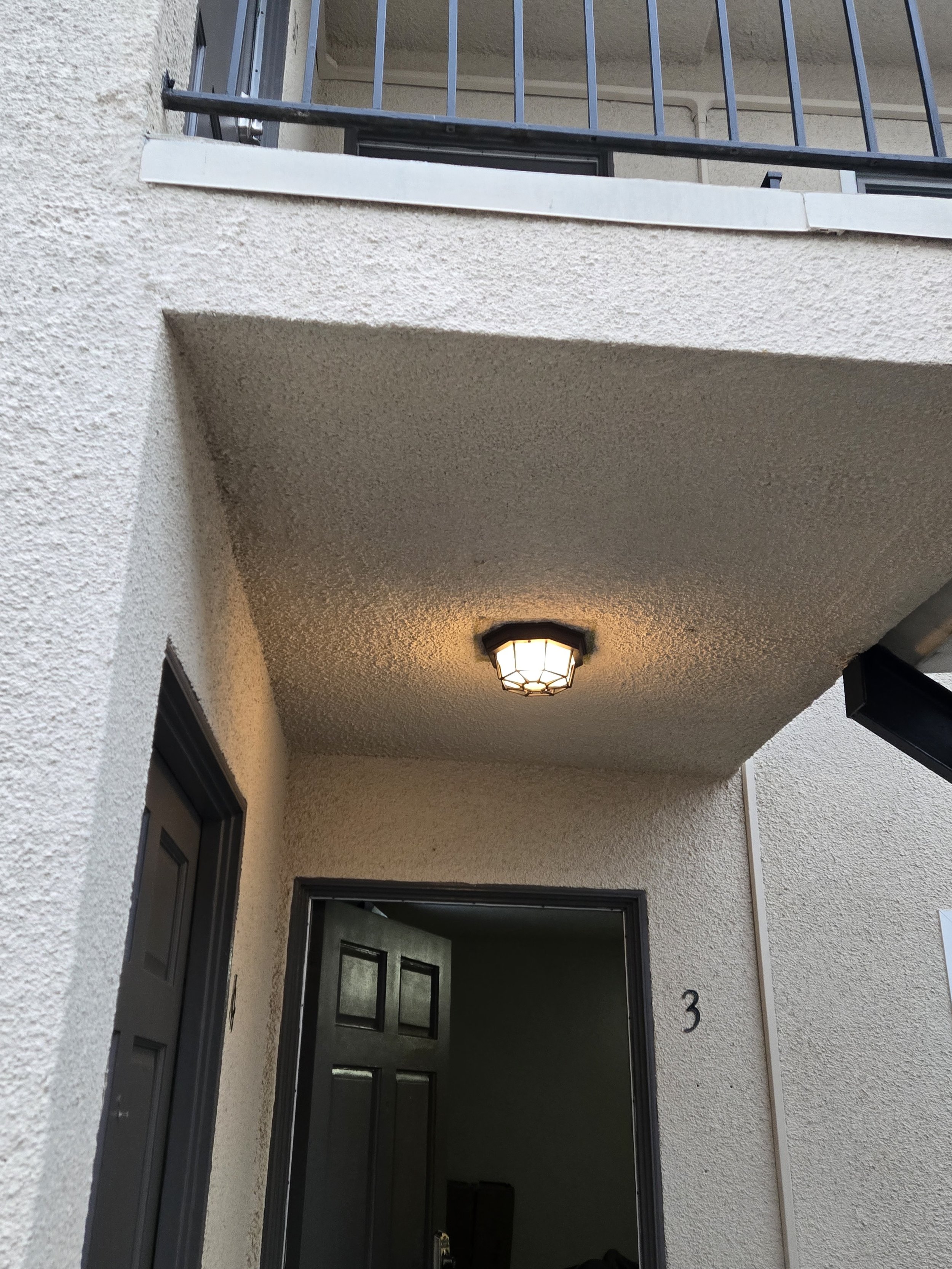 Exterior view of an apartment or condo entrance with a black door and a porch light, including a small balcony with a railing above the entrance.