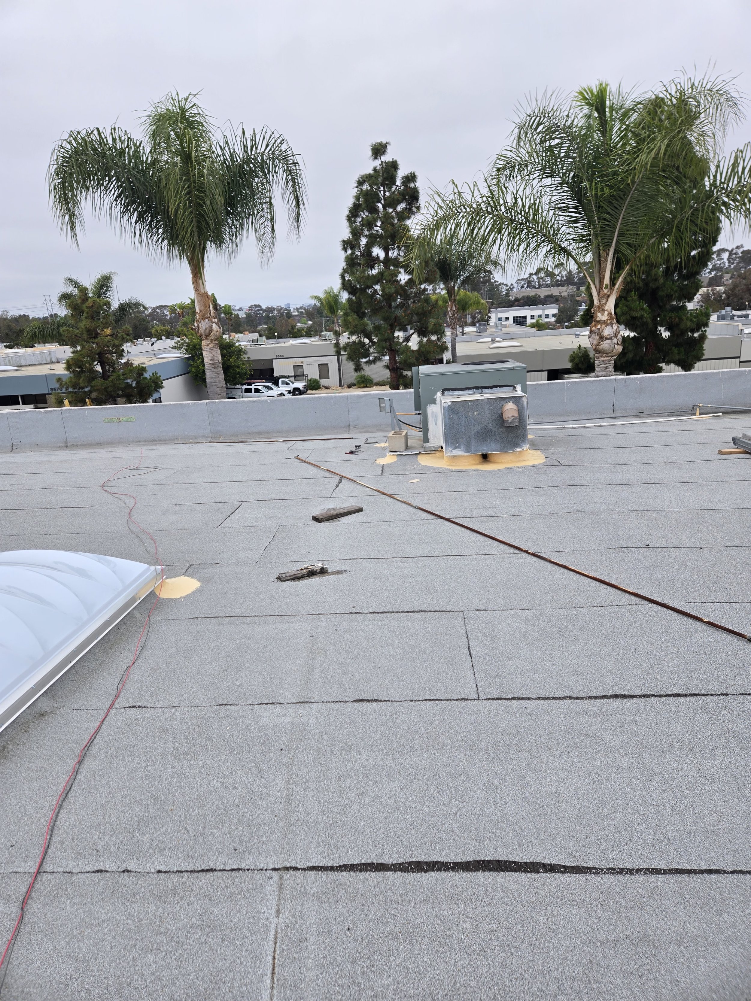 View of a flat rooftop with gray roofing material, an HVAC unit or vent, and electrical cables, with trees and industrial buildings in the background under a cloudy sky.