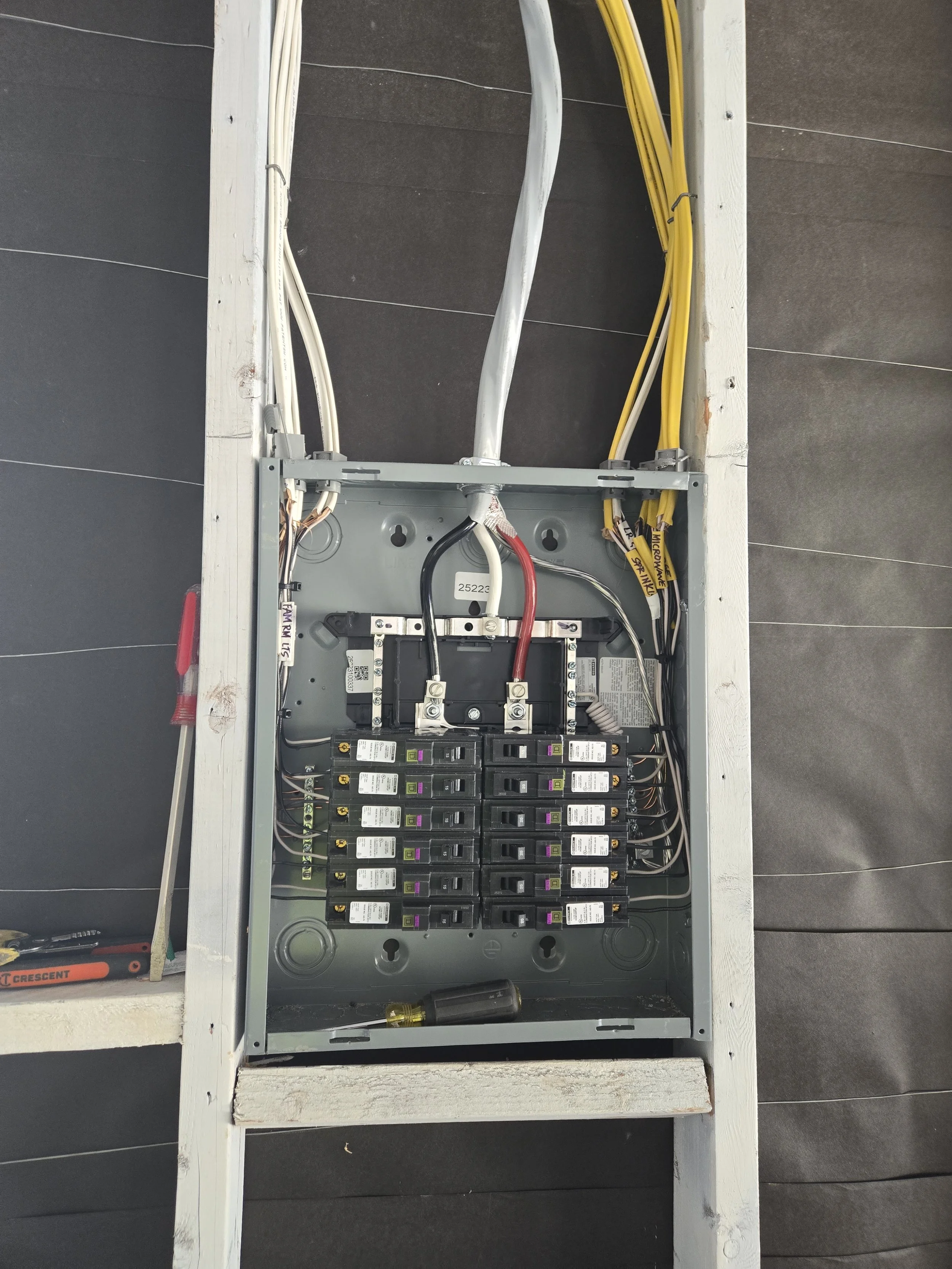 Electrical panel with circuit breakers, wires, and tools mounted on a wooden frame against a black tiled wall.