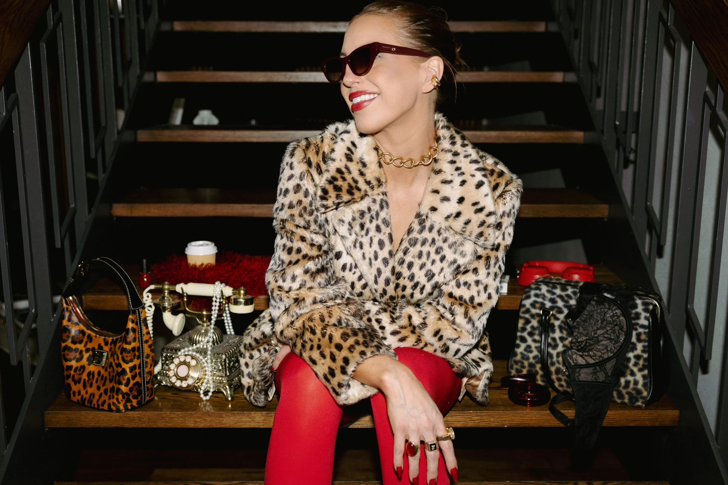 Woman wearing sunglasses and a vintage fur coat, sitting on stairs surrounded by vintage purses and phone.