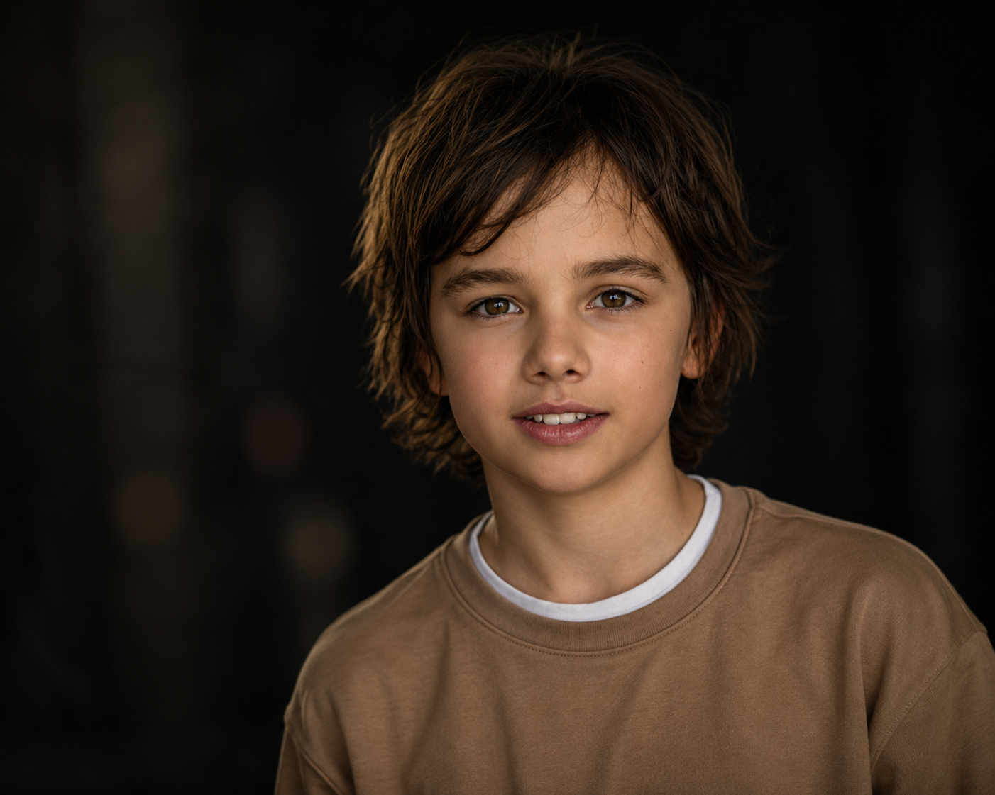 A young boy with wavy brown hair, wearing a beige sweatshirt, looking at the camera with a slight smile, in a dark background.