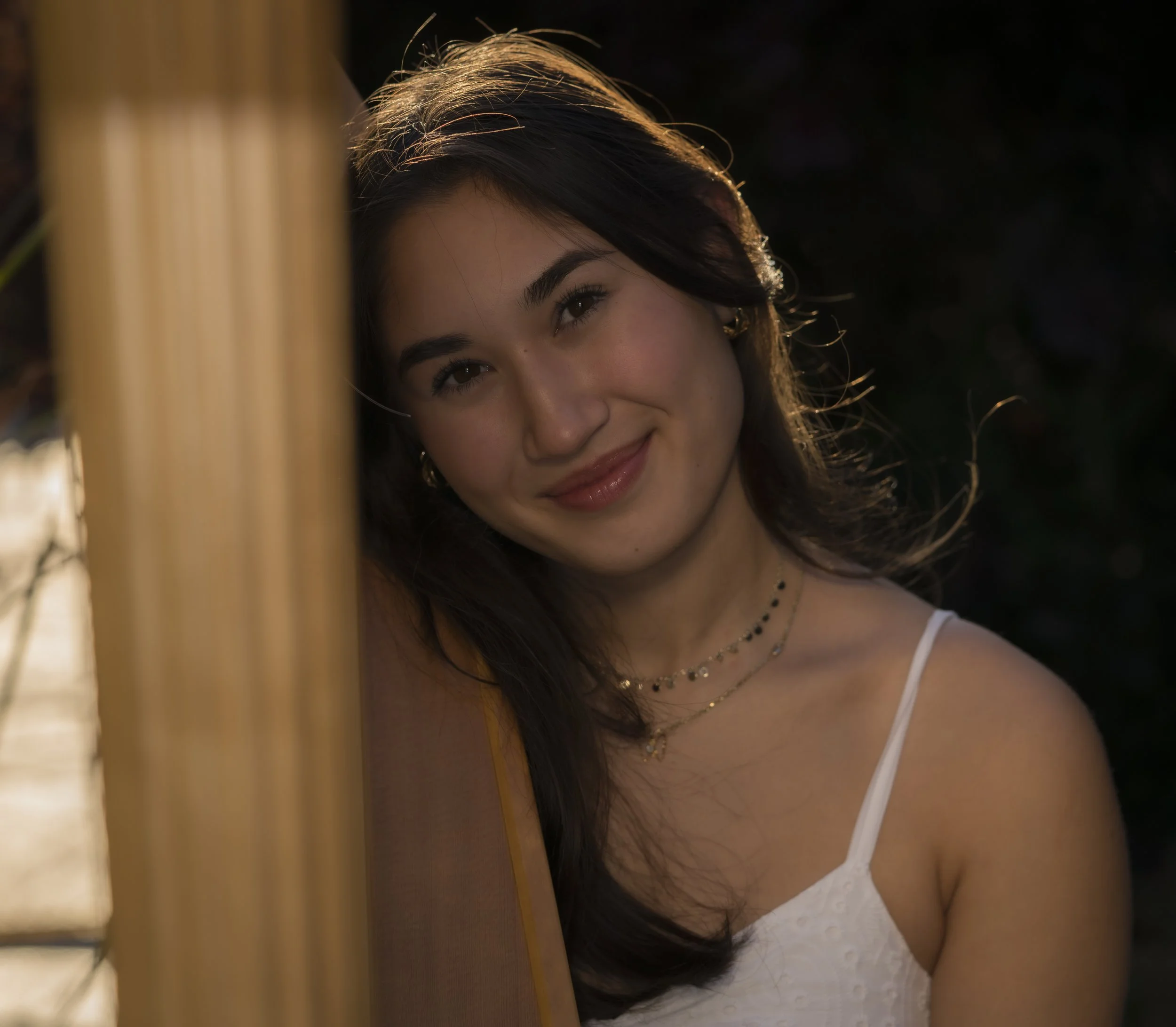 Close-up of a young woman with dark hair and brown eyes, smiling gently and wearing a white top, gold jewelry, and earrings, partially obscured by a vertical wooden object in the foreground, against a dark background with sunlight illuminating her hair.