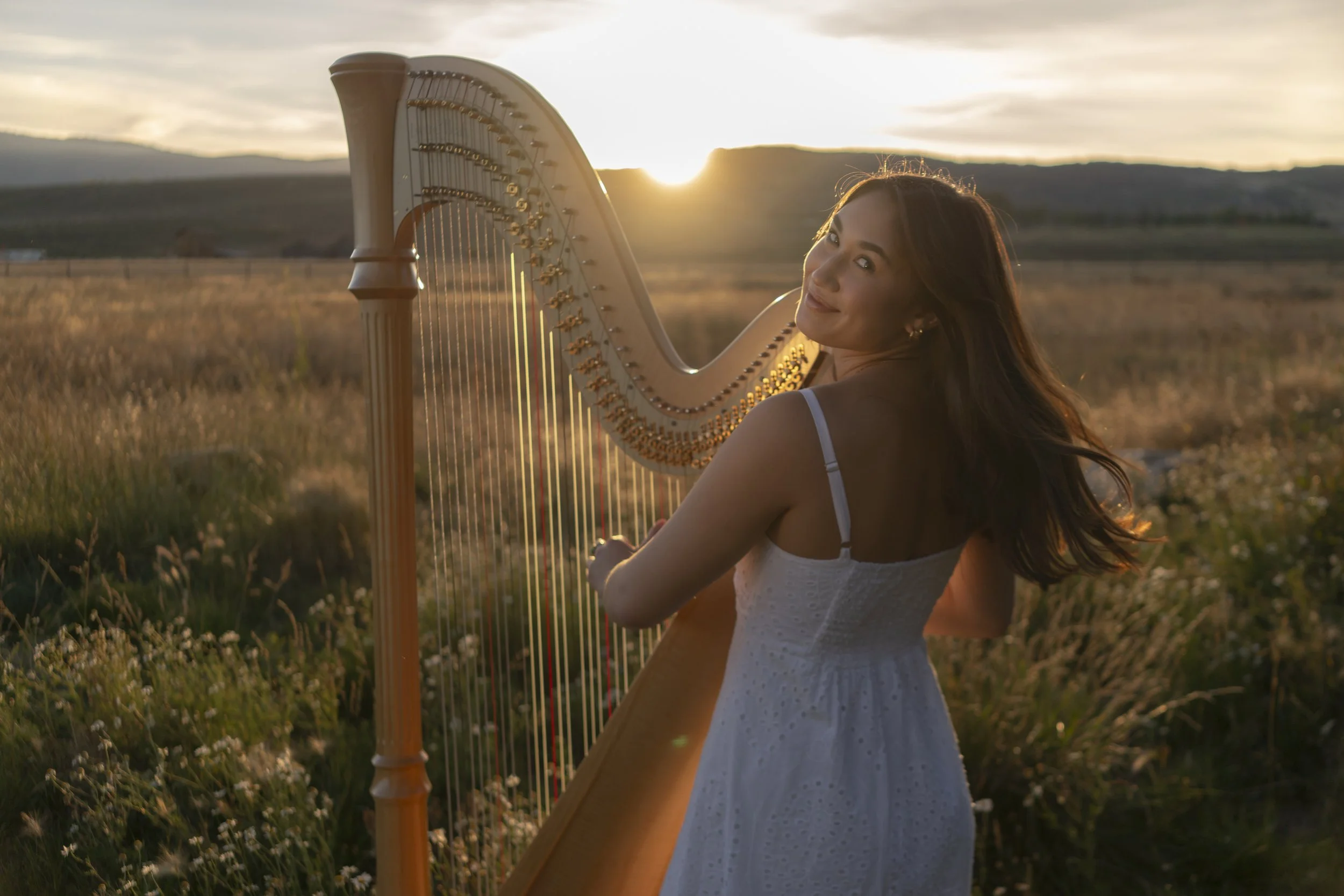 A woman with long brown hair wearing a white dress is smiling and playing a harp outdoors during sunset, in a field of tall grass with mountains in the background.