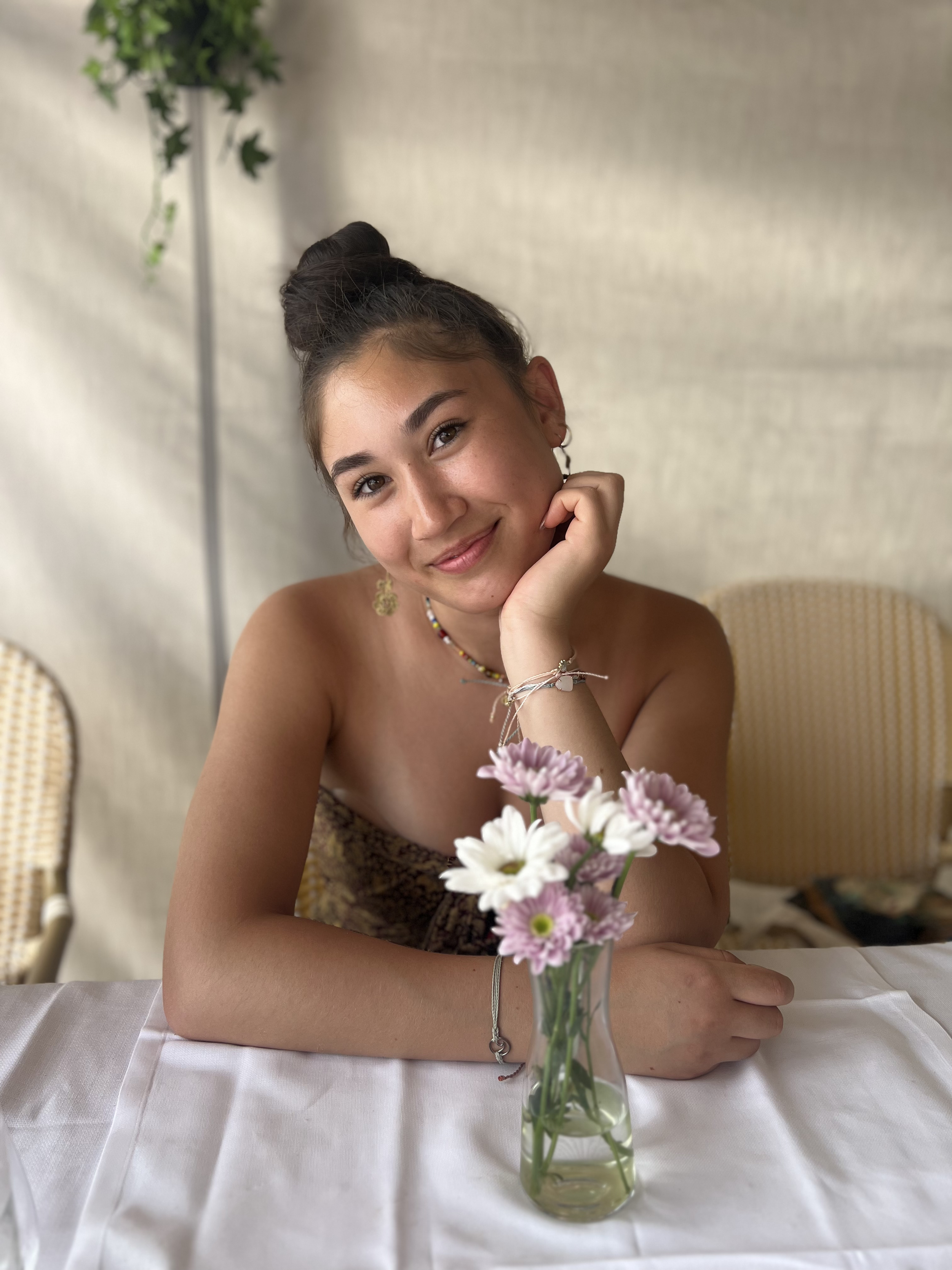 Smiling woman with dark hair in a bun, wearing earrings and necklaces, sitting at a table with a vase of pink and white flowers, resting her chin on her hand.