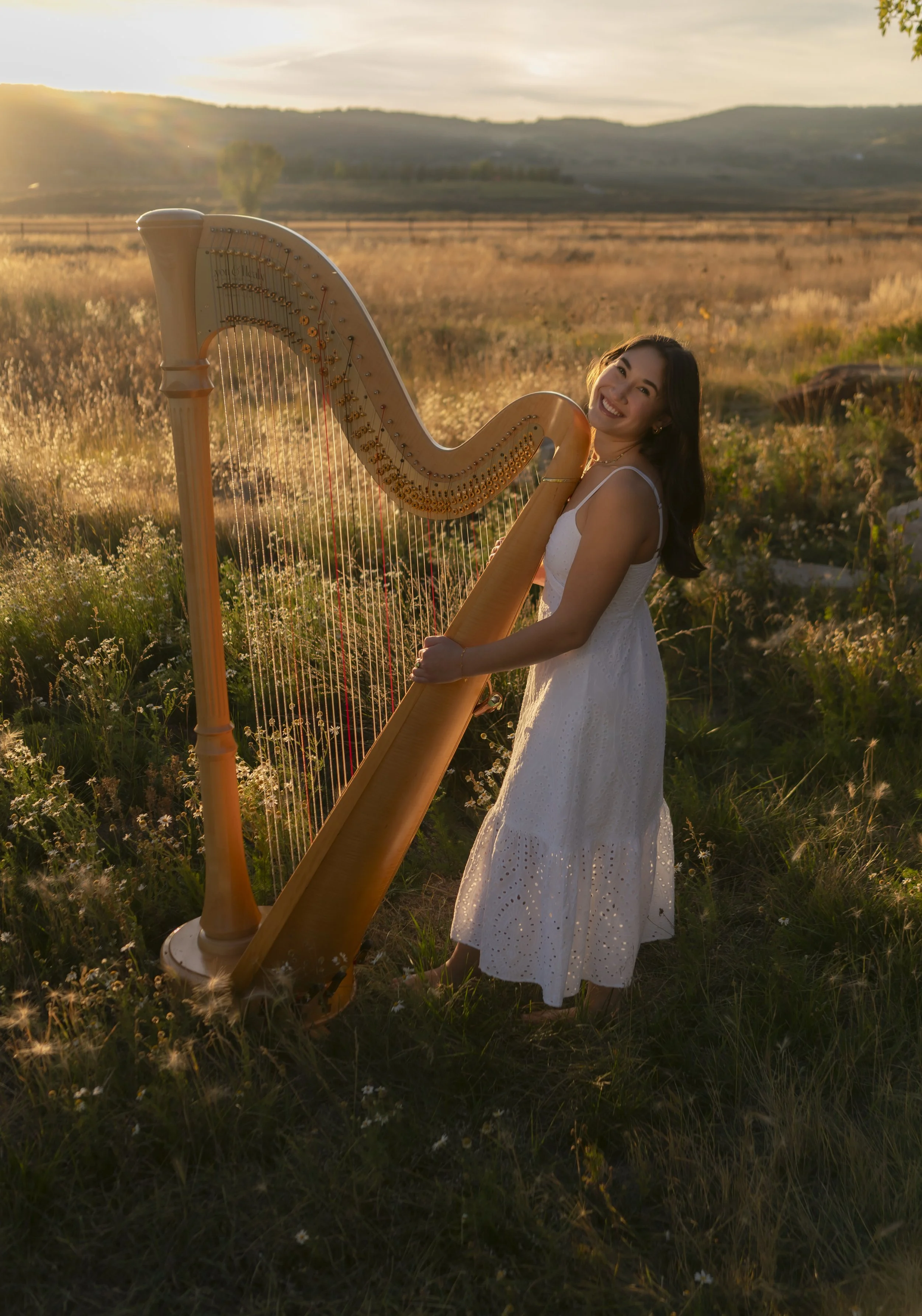 A woman in a white dress smiling while standing in a field at sunset, holding a wooden harp.