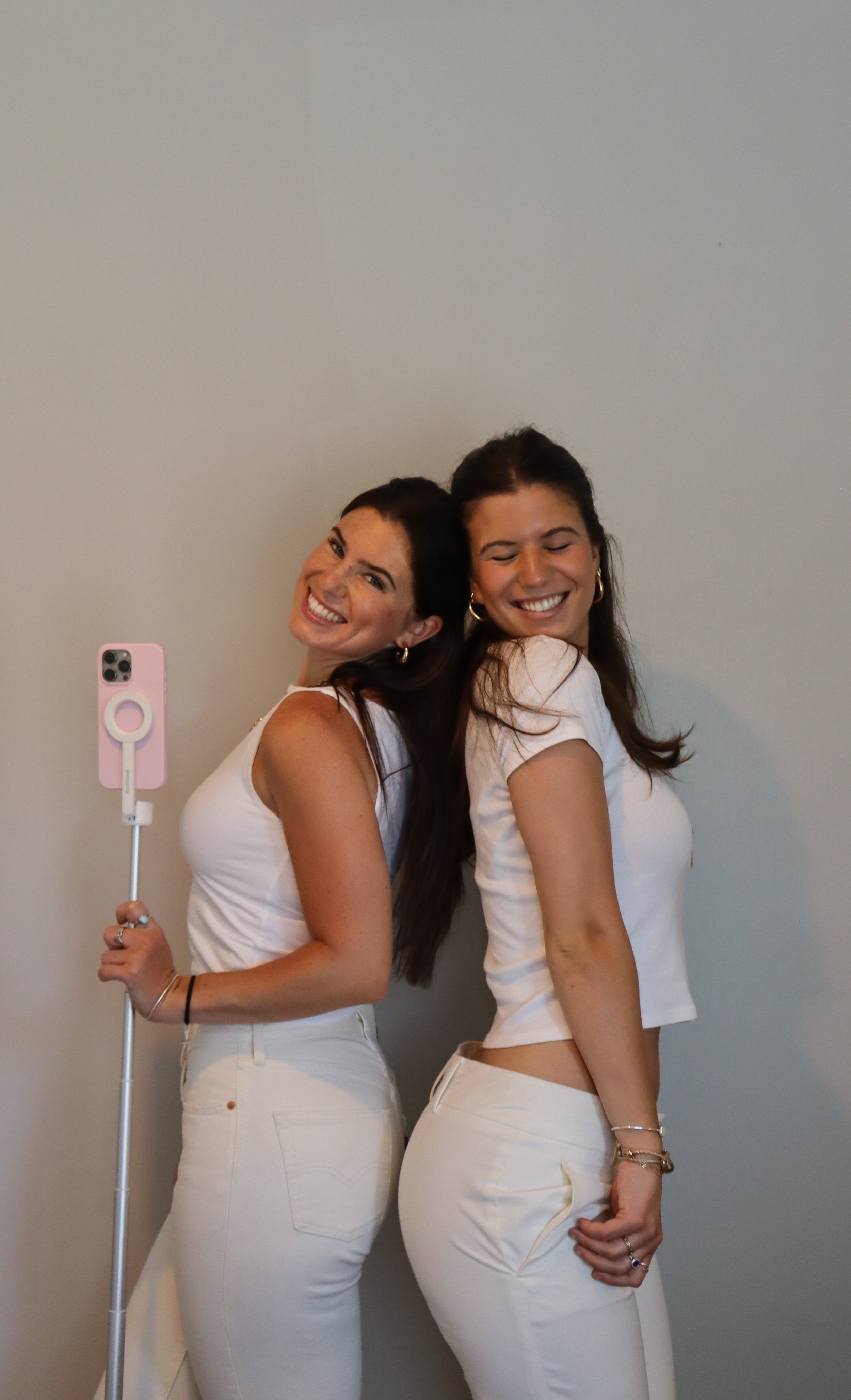 Two women dressed in white, smiling, standing back to back against a plain wall. One holds a pink selfie stick.