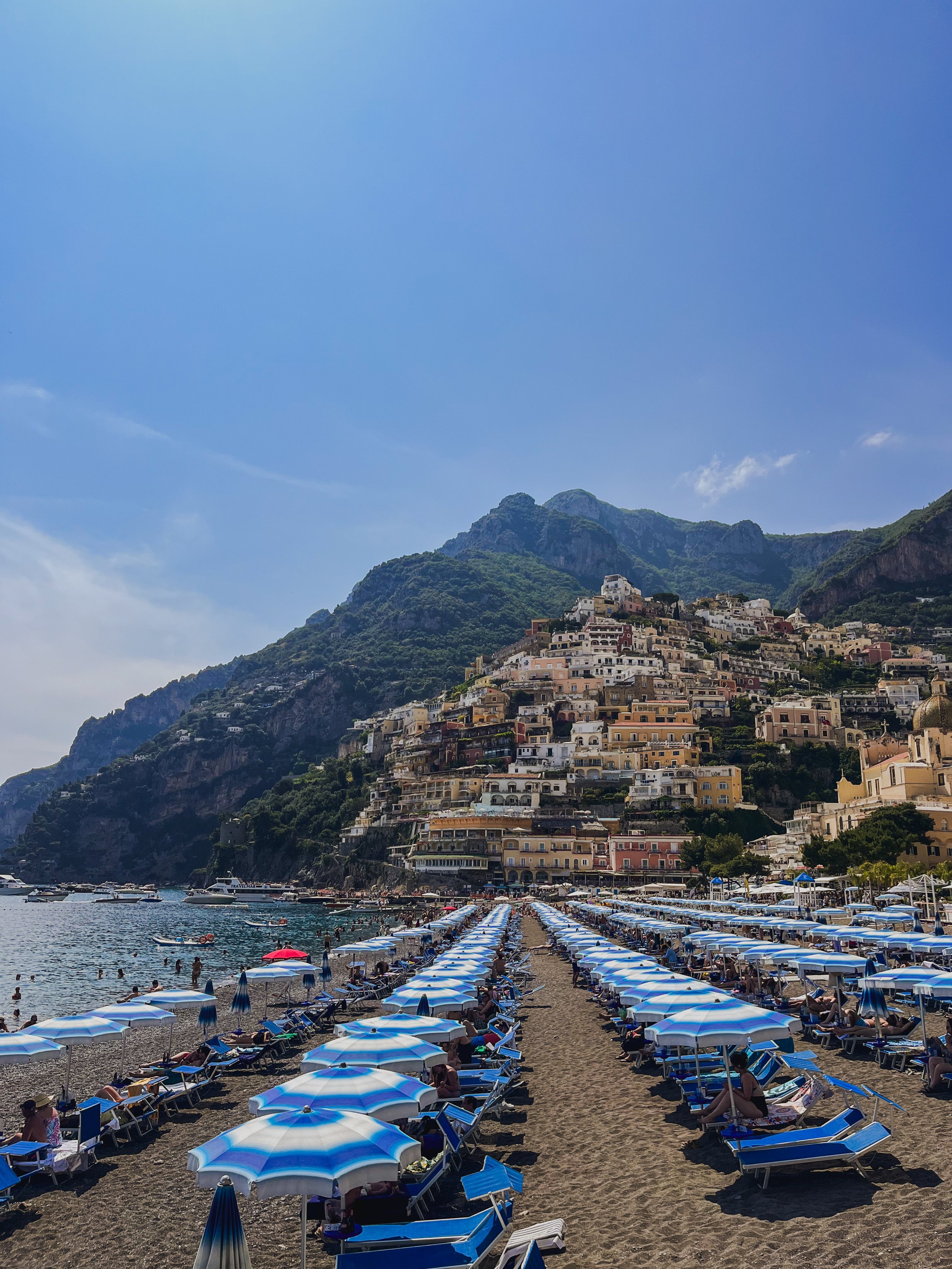 Beach with blue and white umbrellas, sunbathers, and boats docked in the water. Behind the beach, a hillside with colorful buildings rises up toward green mountains under a bright blue sky.