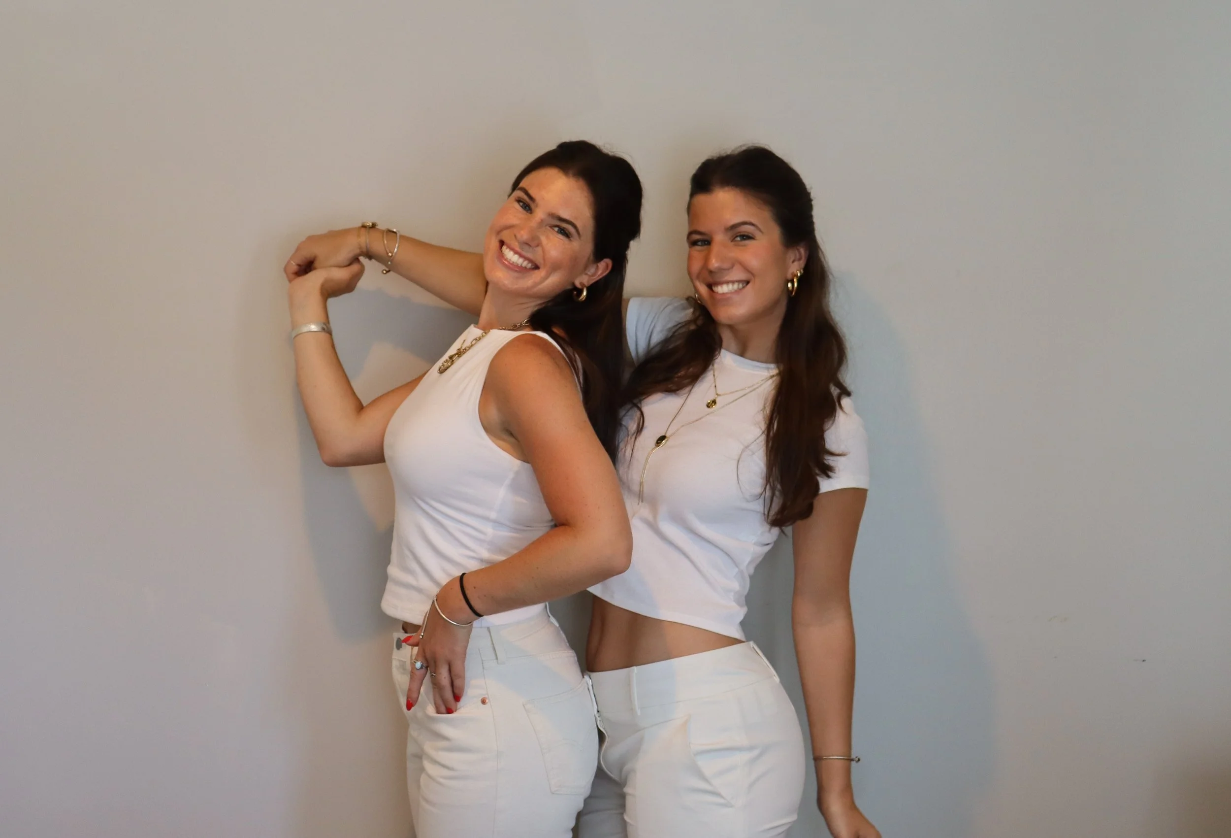 Two smiling women standing close together in front of a plain wall, wearing white tops and pants, with bracelets and jewelry.
