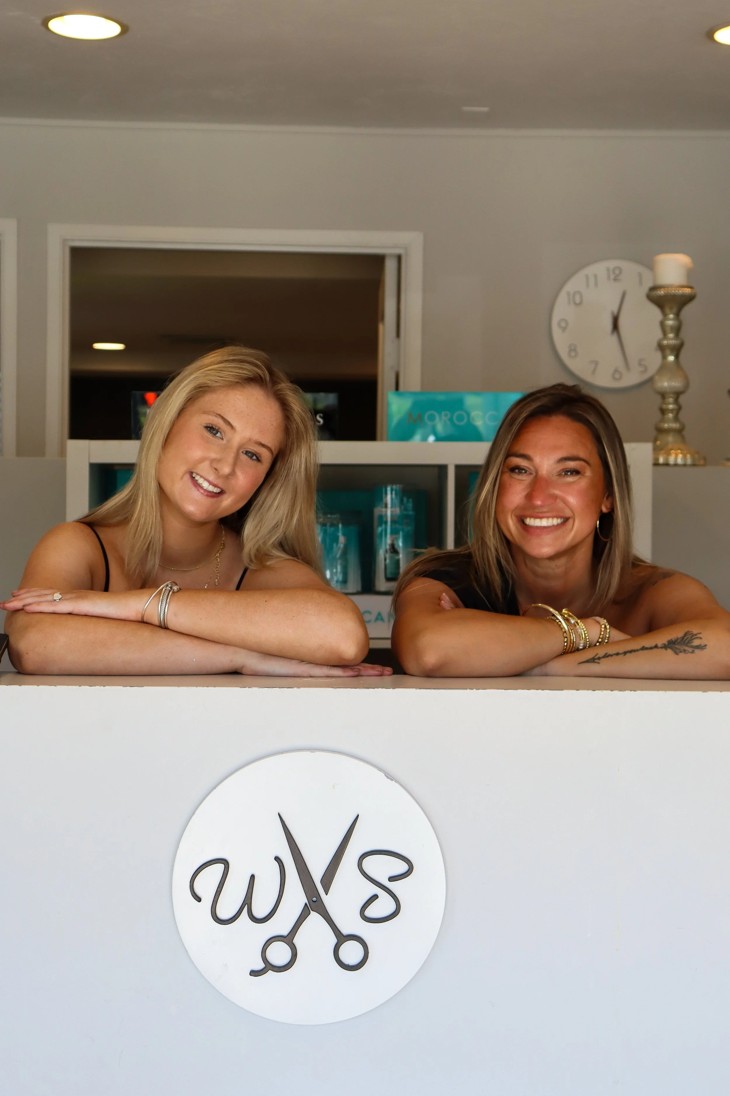Two smiling women with blonde and brunette hair, wearing bracelets, leaning on a white counter with the salon logo, in a modern salon interior with a clock and candles in the background.