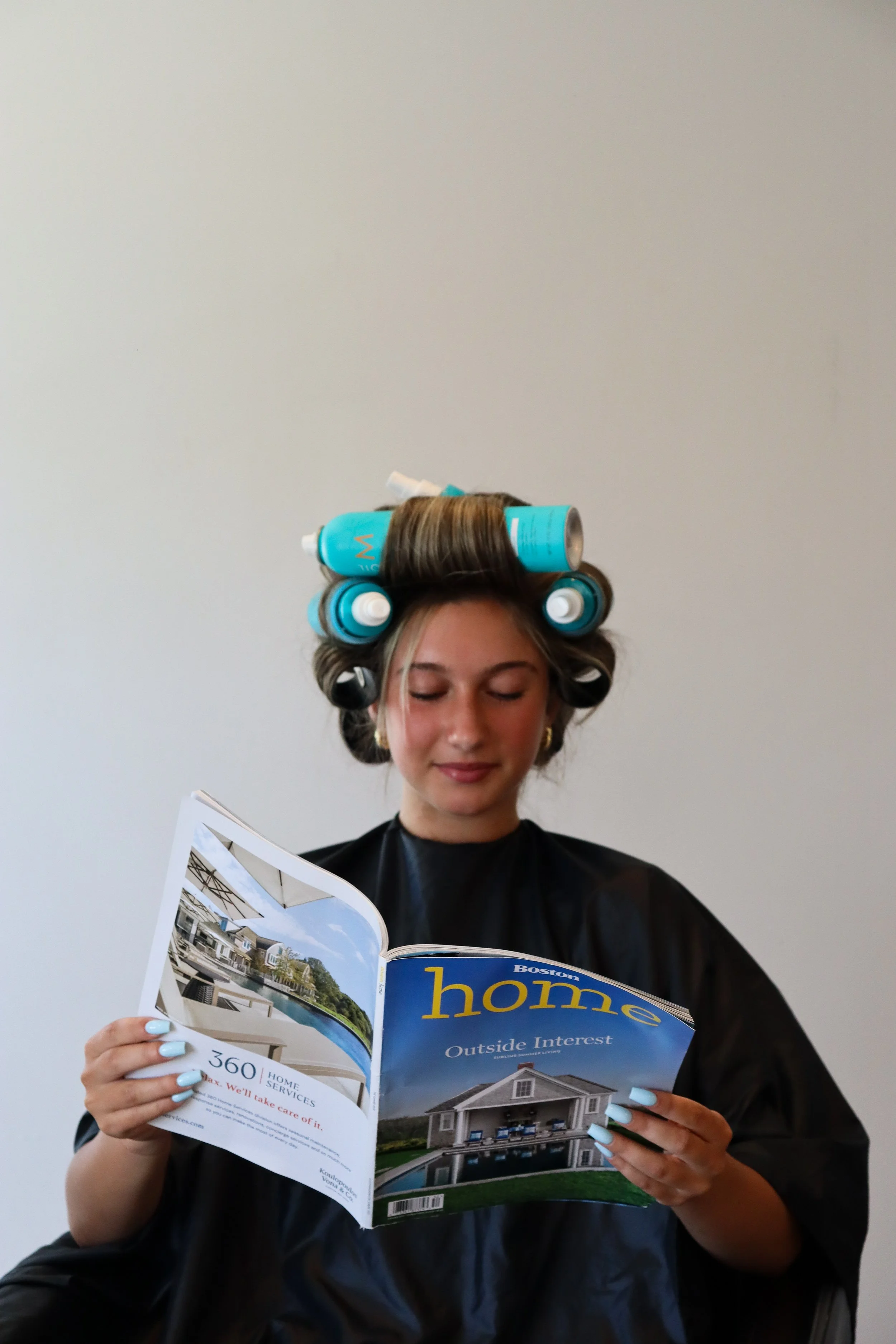 A young woman with curlers in her hair, reading a magazine titled 'Boston Home' with a picture of a house, against a plain white wall.